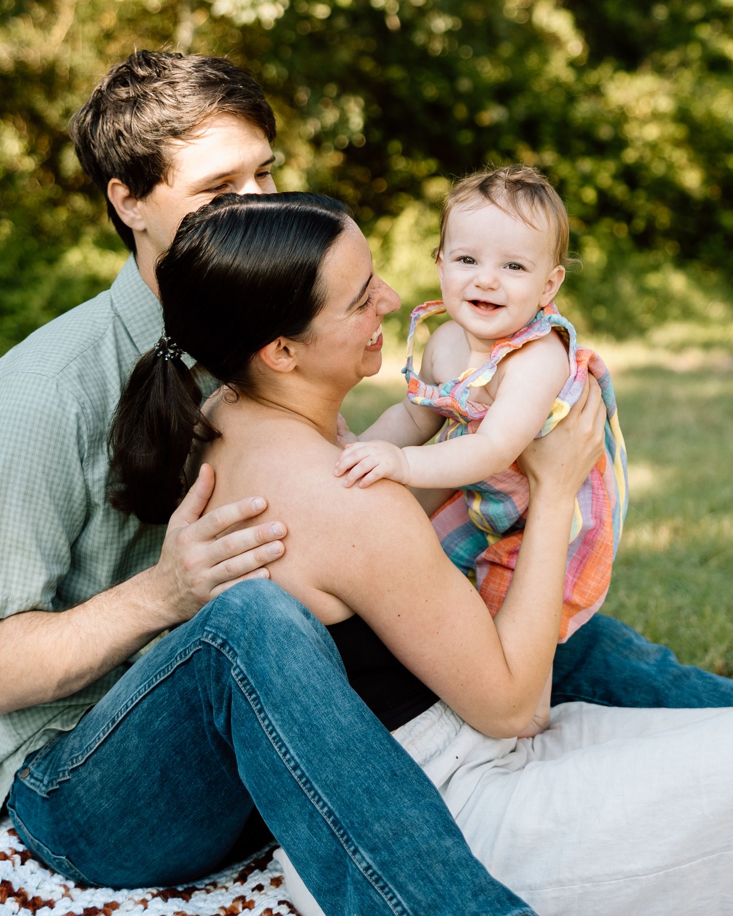 A happy family of three sits outdoors on grass, parents smiling at their joyful baby girl in a colorful dress.
