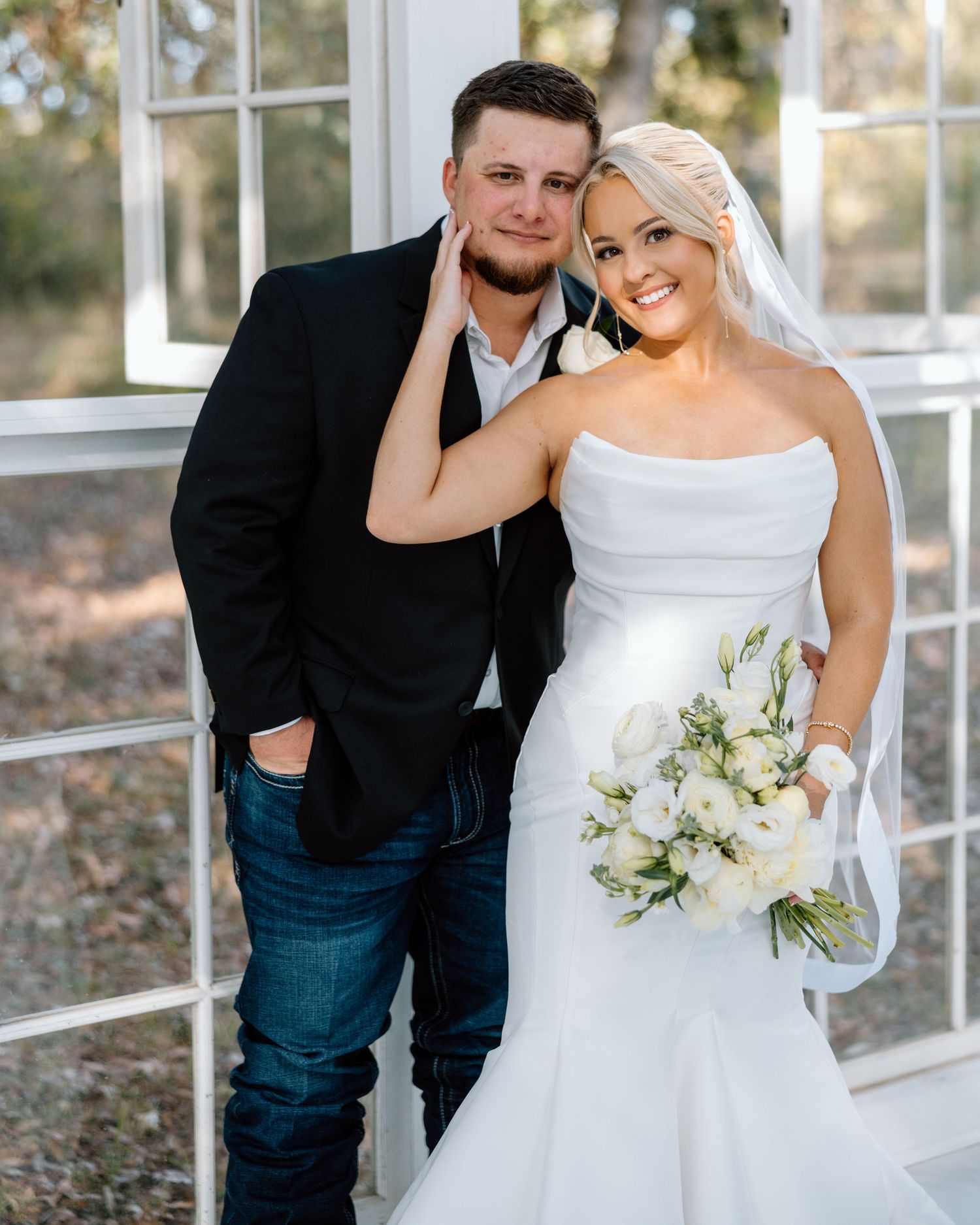 Smiling bride in strapless mermaid gown holding white bouquet poses with groom in black blazer.