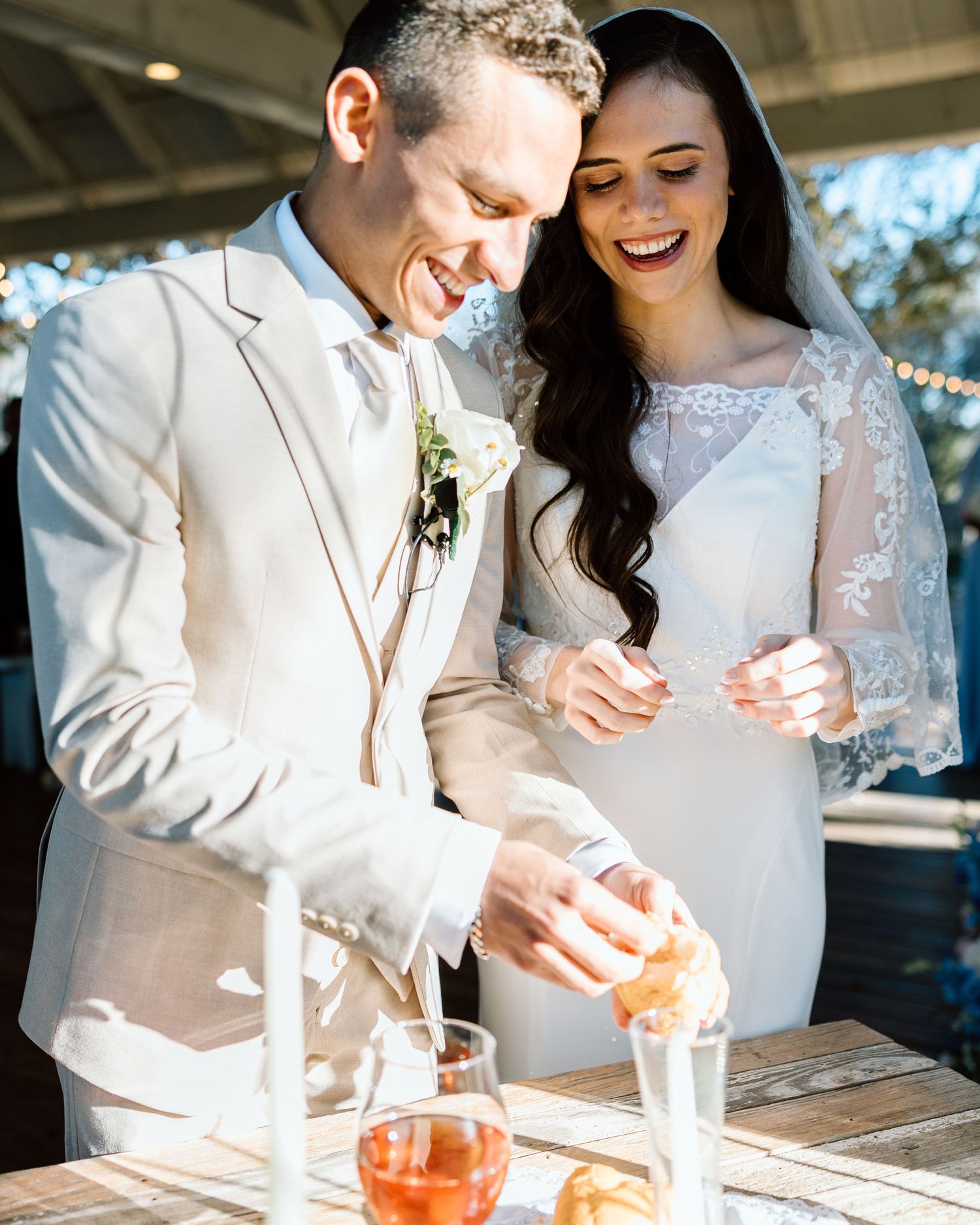 Happy bride and groom cutting wedding cake together outdoors, bride in lace gown, groom in beige suit.