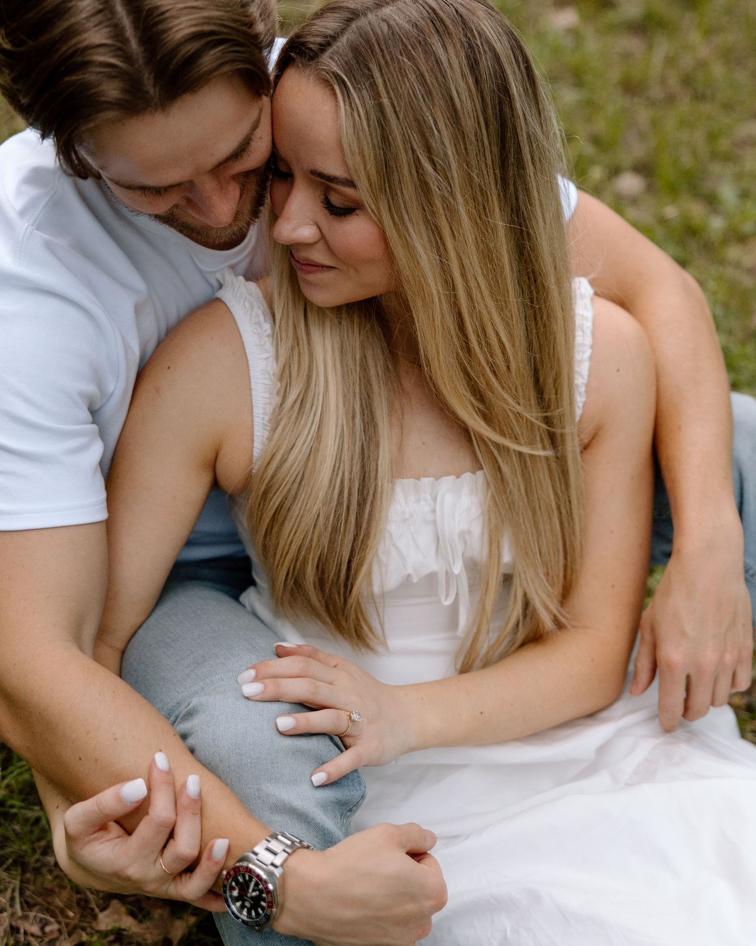 Romantic couple sitting together on grass, woman in white dress leaning into man wearing a watch.