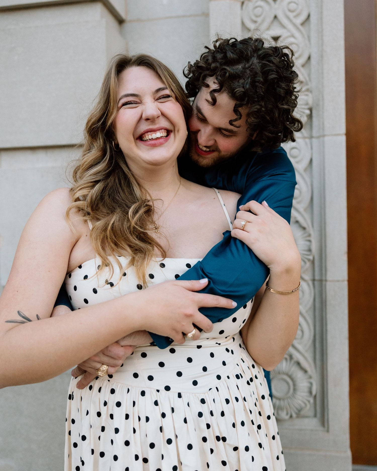 Laughing couple embracing near ornate building, woman in polka dot dress and man in teal shirt.