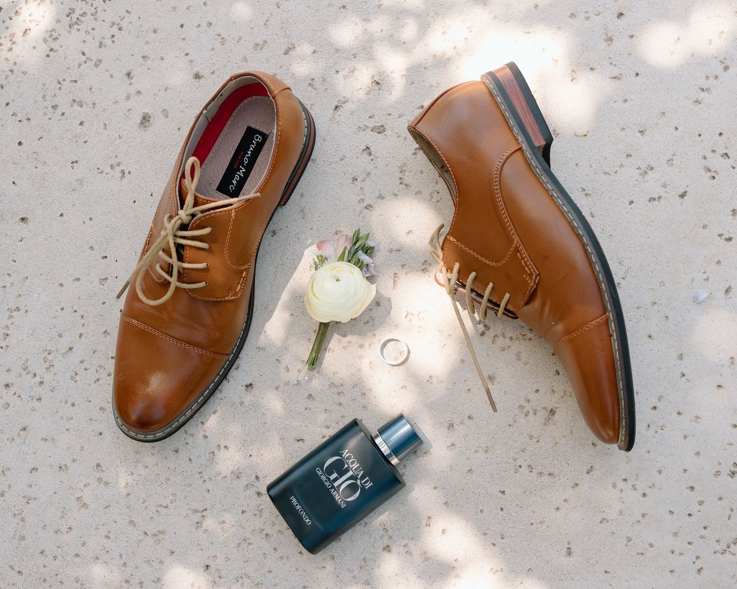 Brown leather dress shoes arranged with a white boutonniere and cologne bottle on a light surface.