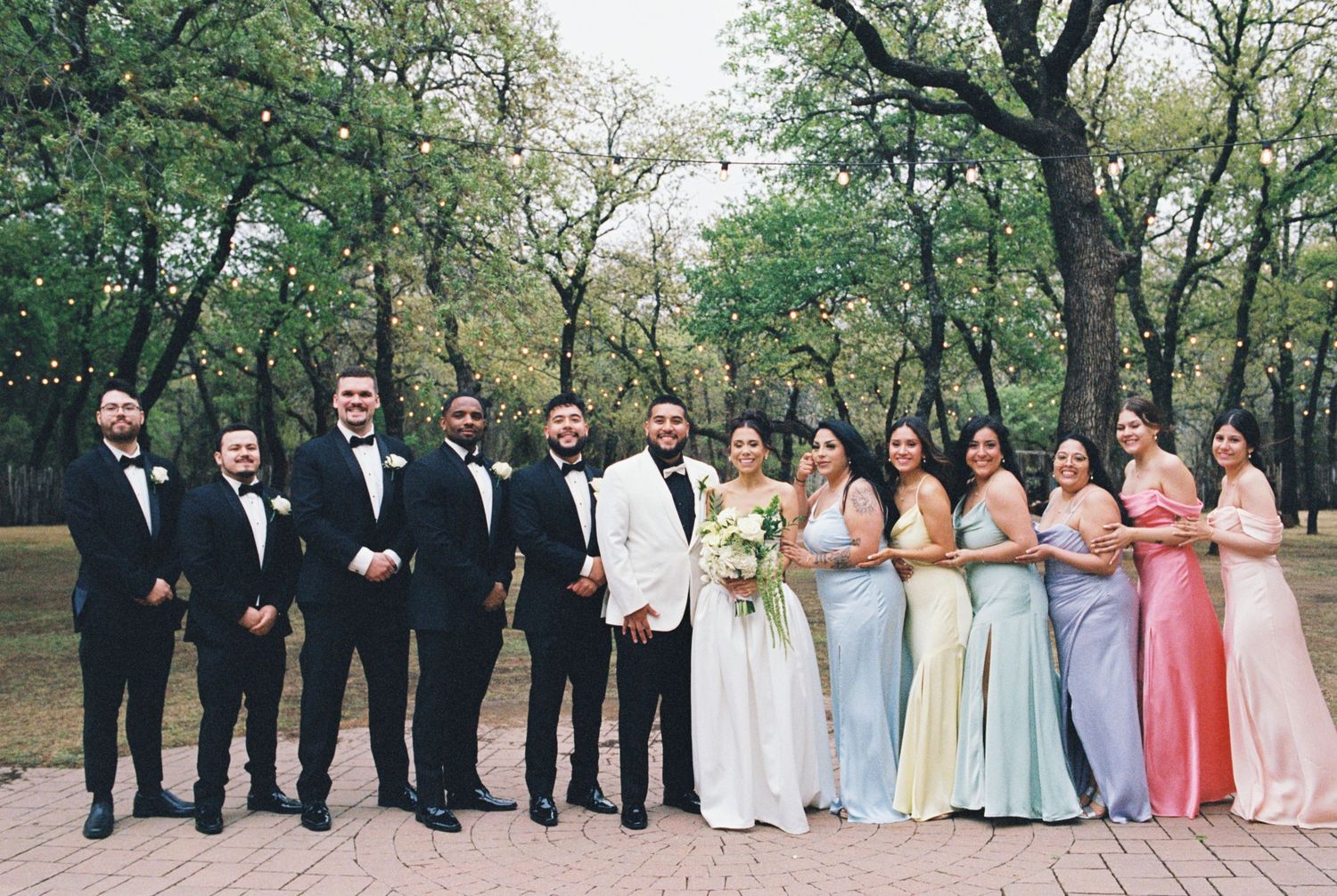 Wedding party in formal attire poses outdoors with groomsmen in black and bridesmaids in pastels.