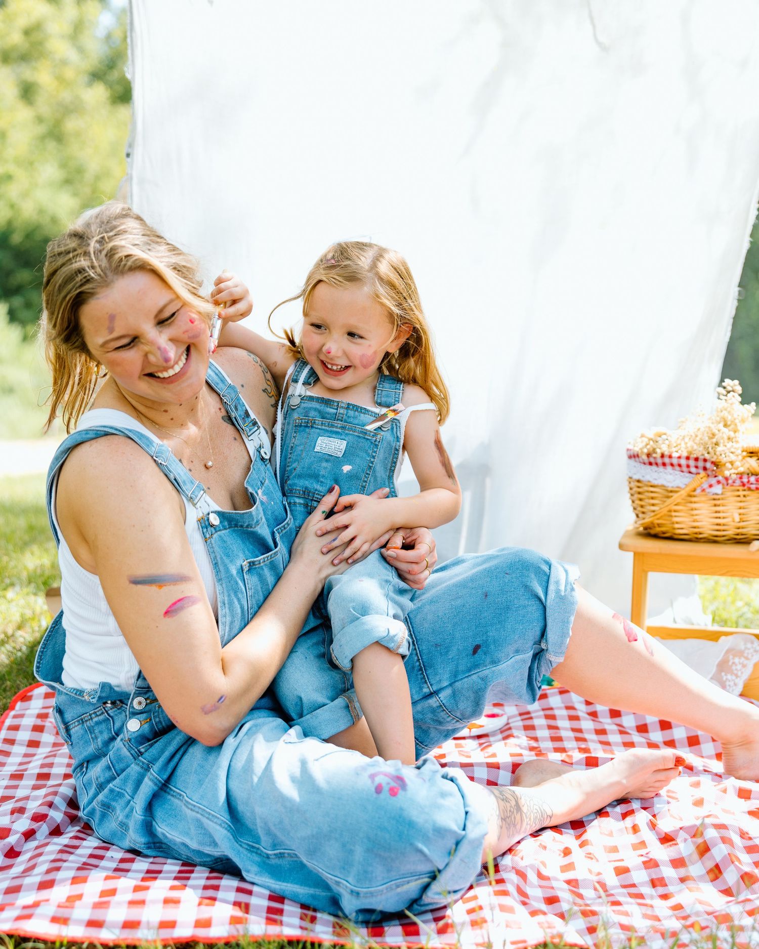 Woman and young girl in matching denim overalls laughing on a red checkered picnic blanket outdoors.