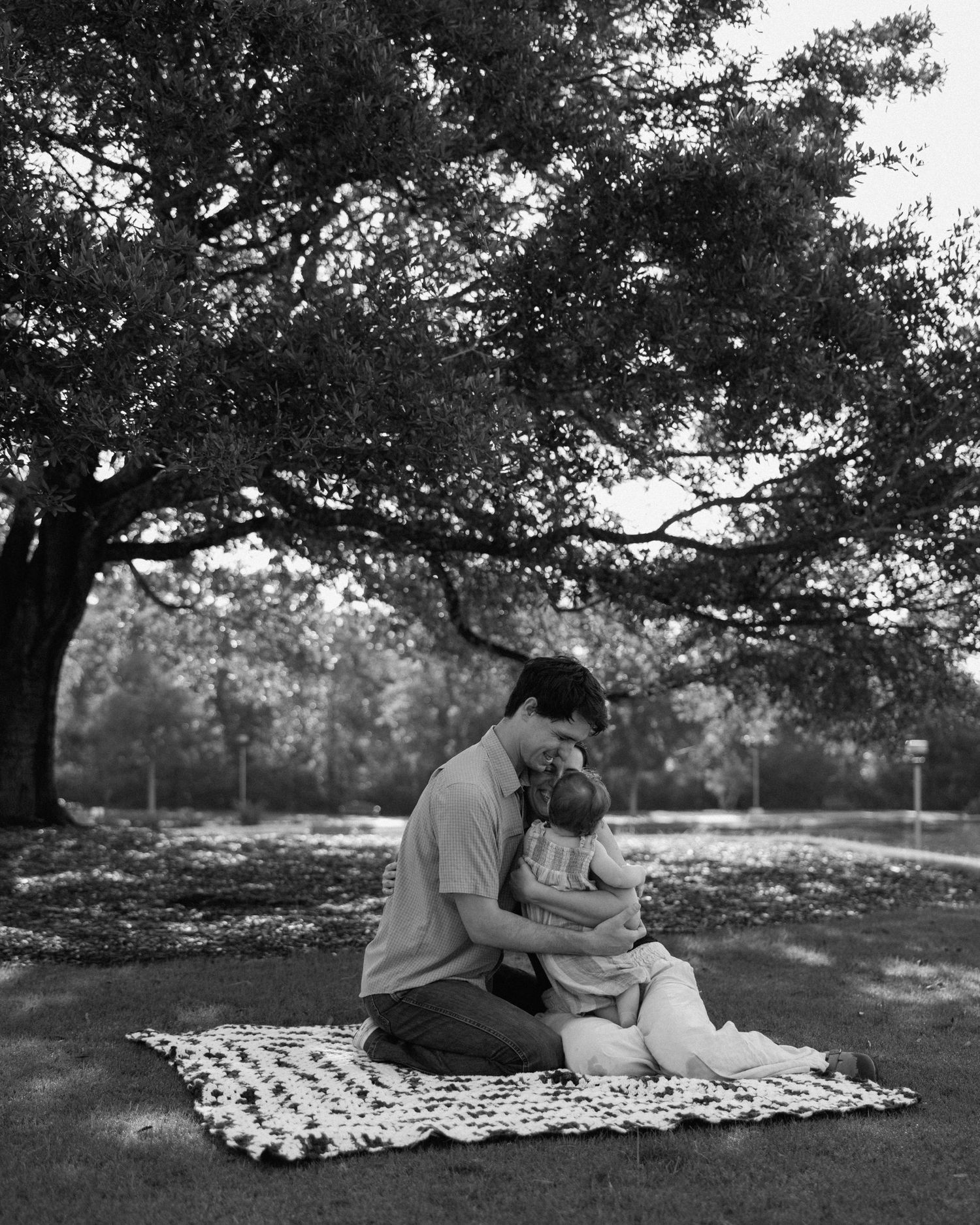 Black and white photo of a parent cradling a baby on a blanket under a large tree in a park.