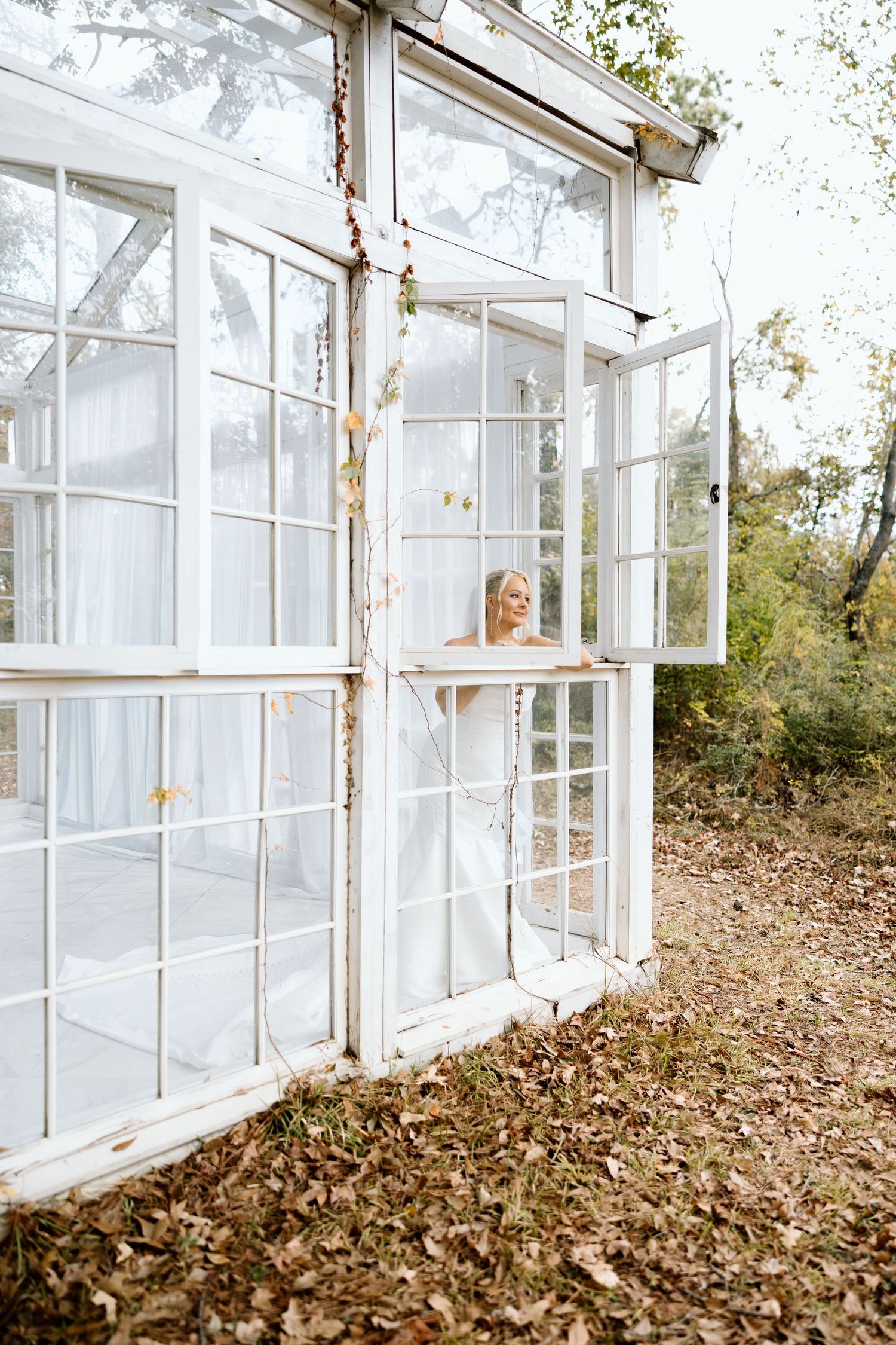 Bride in white wedding gown standing inside a rustic white greenhouse surrounded by autumn foliage.