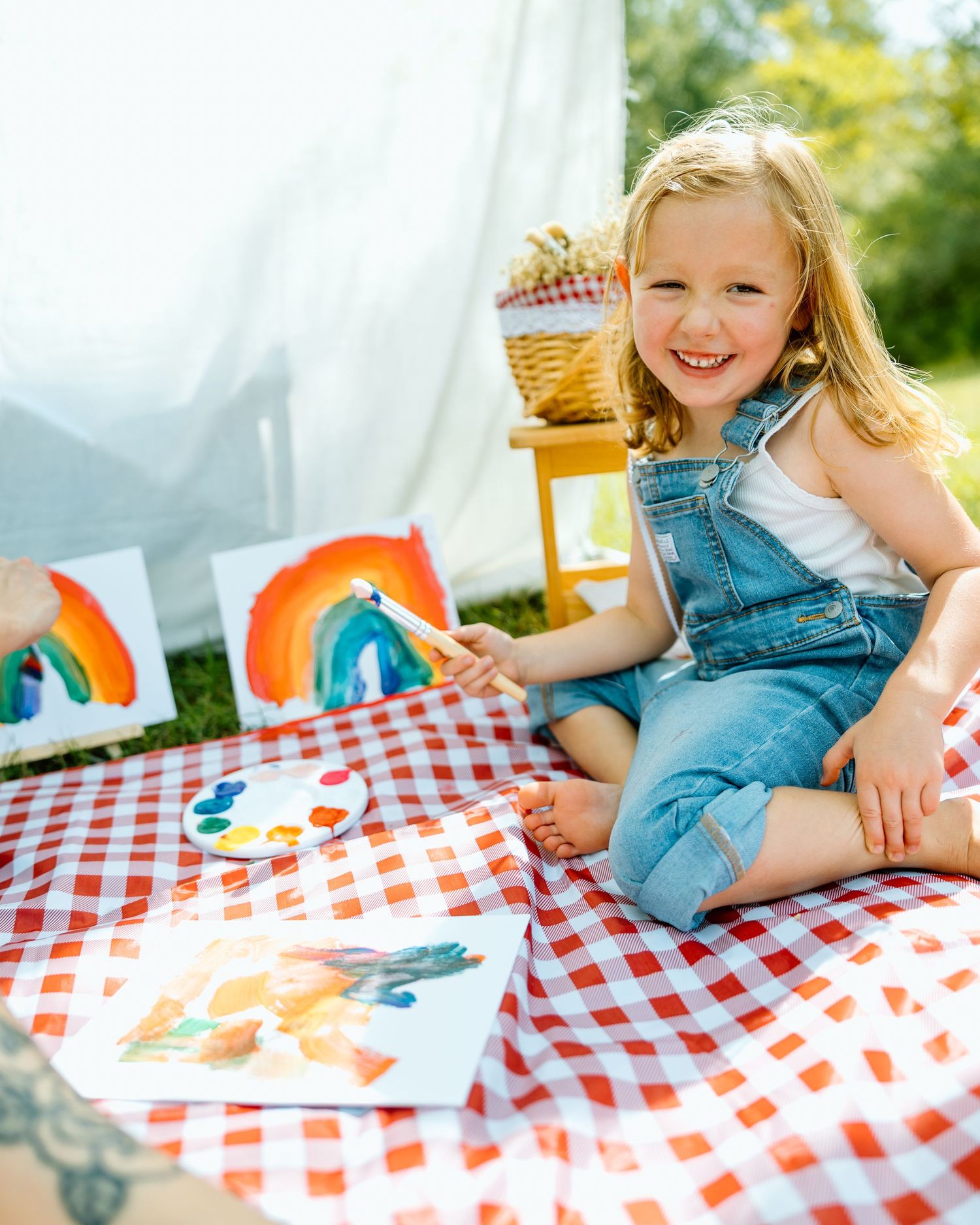 A smiling young girl in denim overalls painting rainbows on canvas while sitting on a red gingham picnic blanket outdoors.