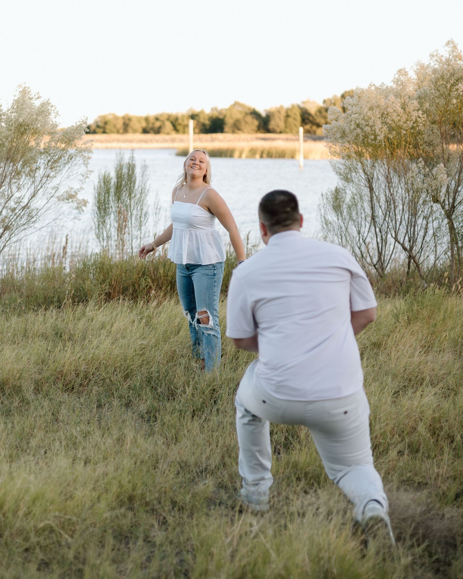Man kneeling in grass proposing to a smiling woman in white top and jeans near a scenic lakeside.