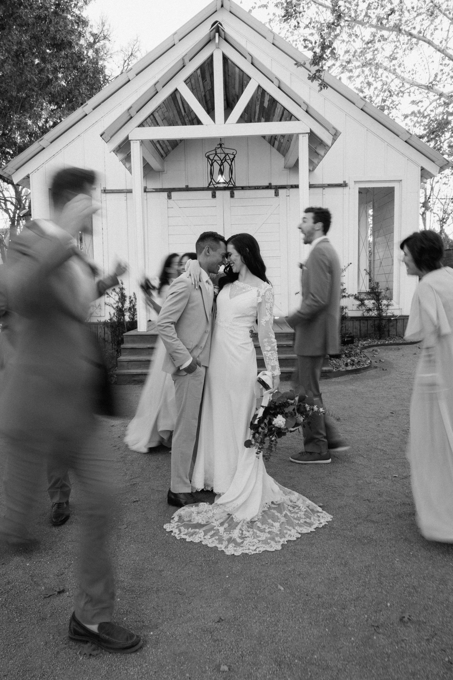 Black and white photo of bride and groom kissing outside a white chapel surrounded by wedding guests.