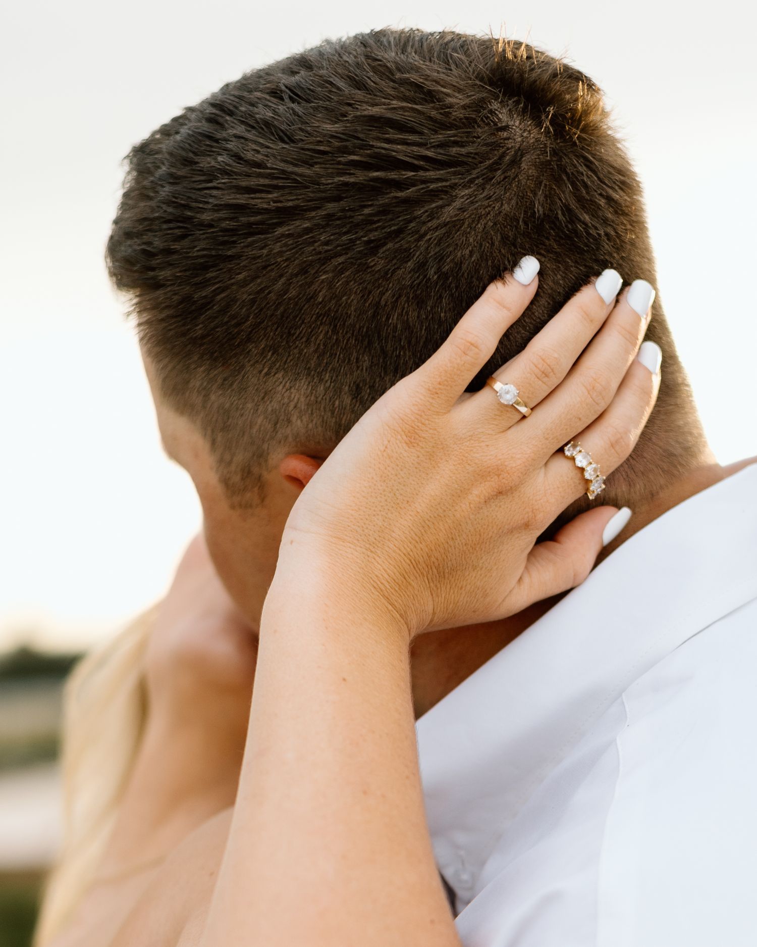 Close-up of a woman's hand with engagement and wedding rings resting on a man's head during a romantic embrace.
