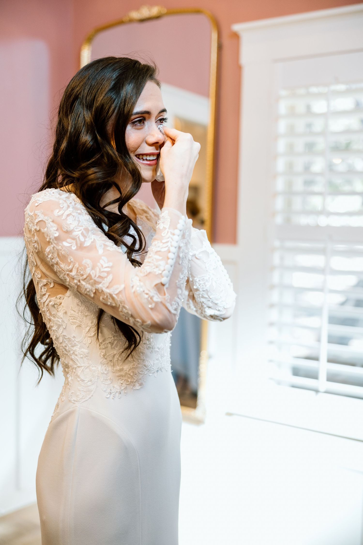 Joyful bride in lace long-sleeve wedding gown laughing while getting ready in pink bridal suite.