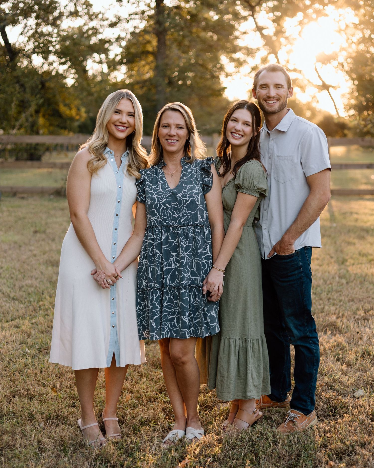Four smiling family members pose together in a sunny outdoor field during golden hour, dressed in casual summer attire.