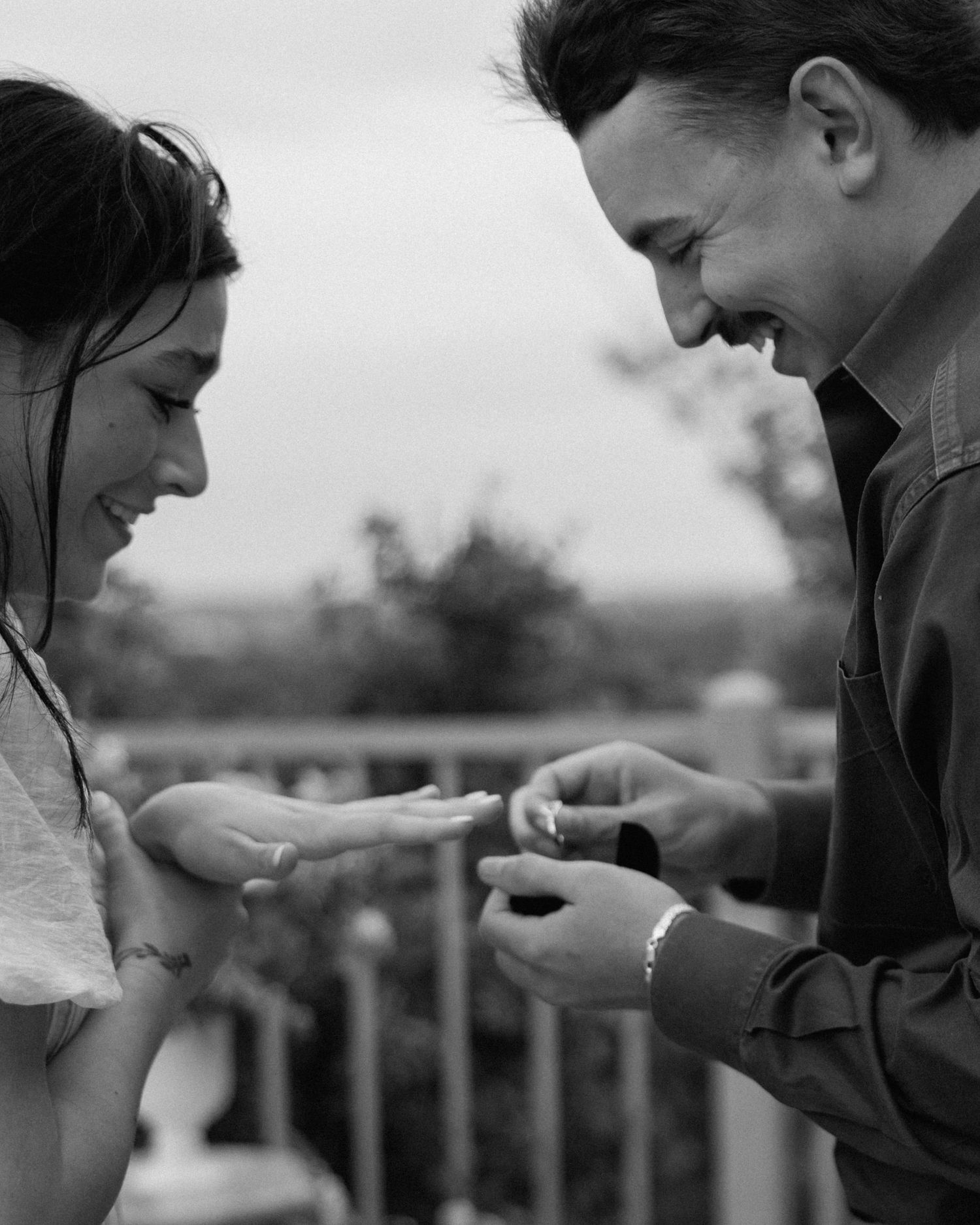 Black and white photo of a couple smiling as a man places a ring on a womans hand during a proposal.