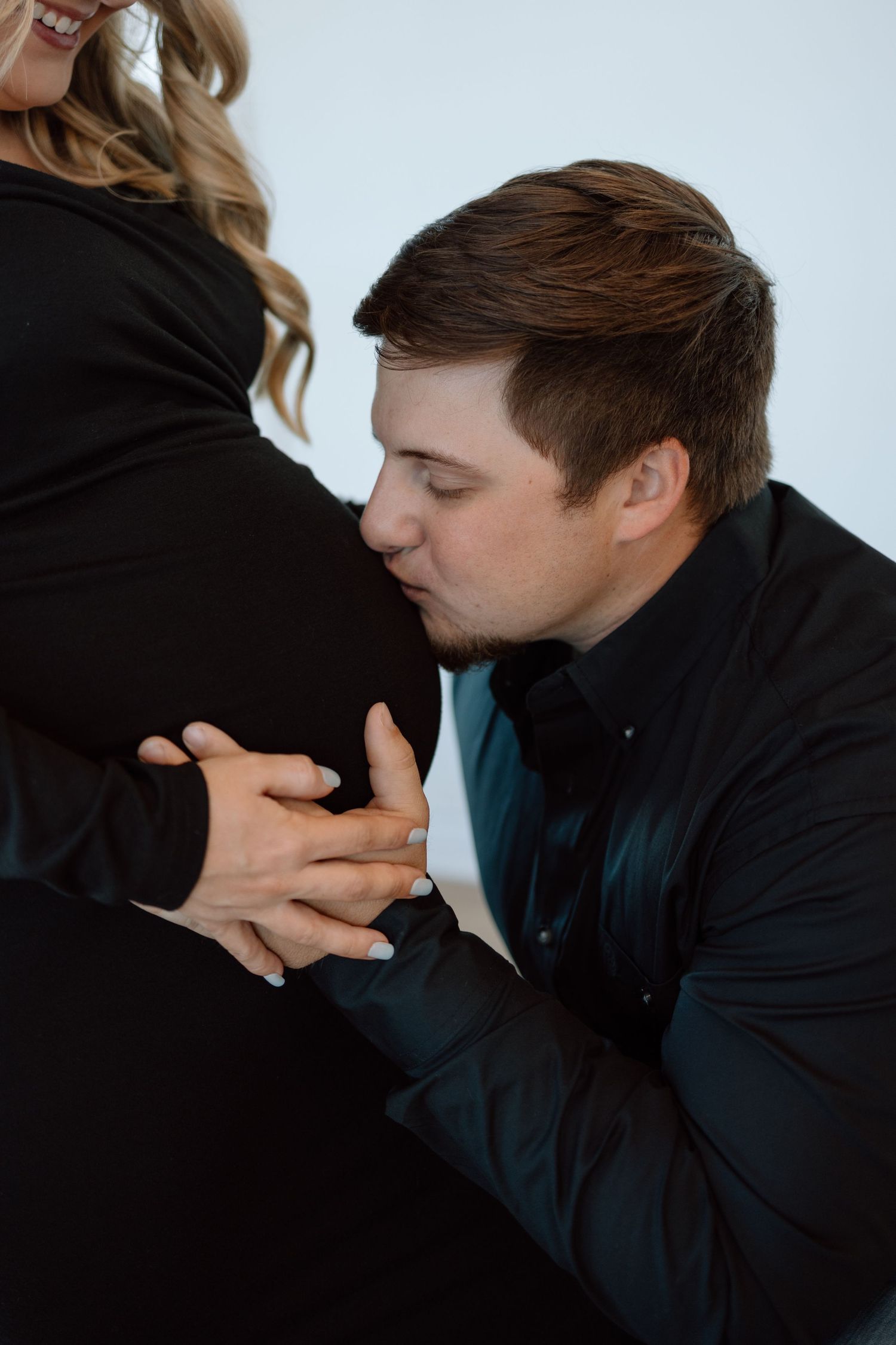 A tender pregnancy photo with someone kissing a baby bump while holding hands, wearing a black and teal outfit.