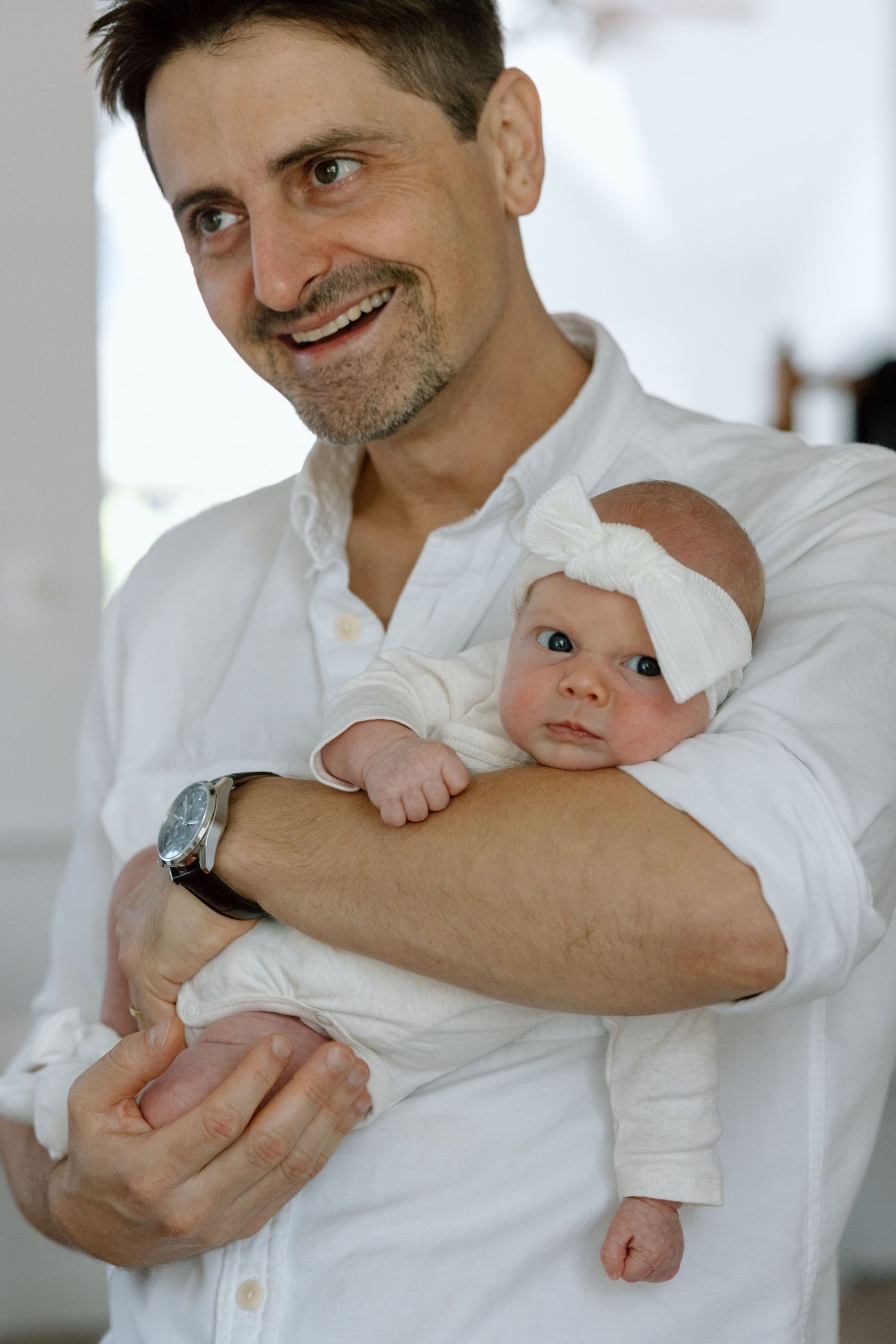 Baby in white outfit with bow headband being held by parent in crisp white button-down shirt.