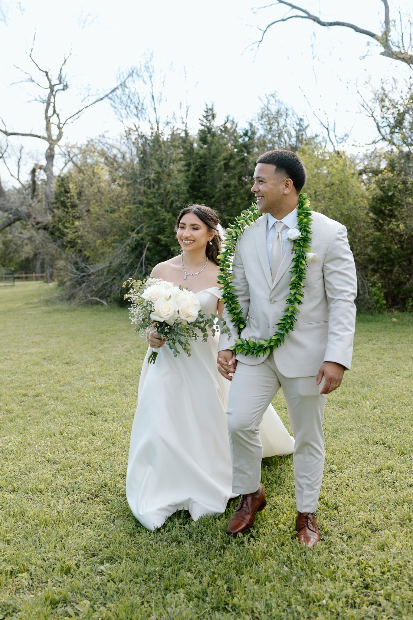 Newlyweds walk hand in hand across lawn wearing traditional white wedding attire and Hawaiian lei.