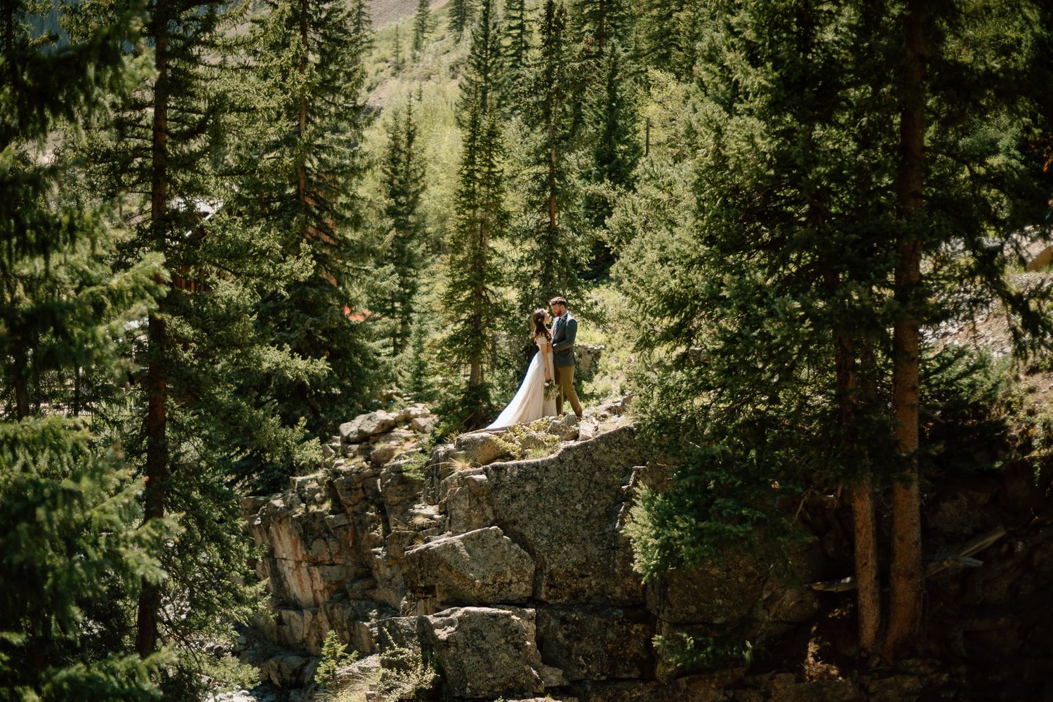 A scenic mountain wedding photo with figures standing on rocky outcrop among tall evergreen trees.