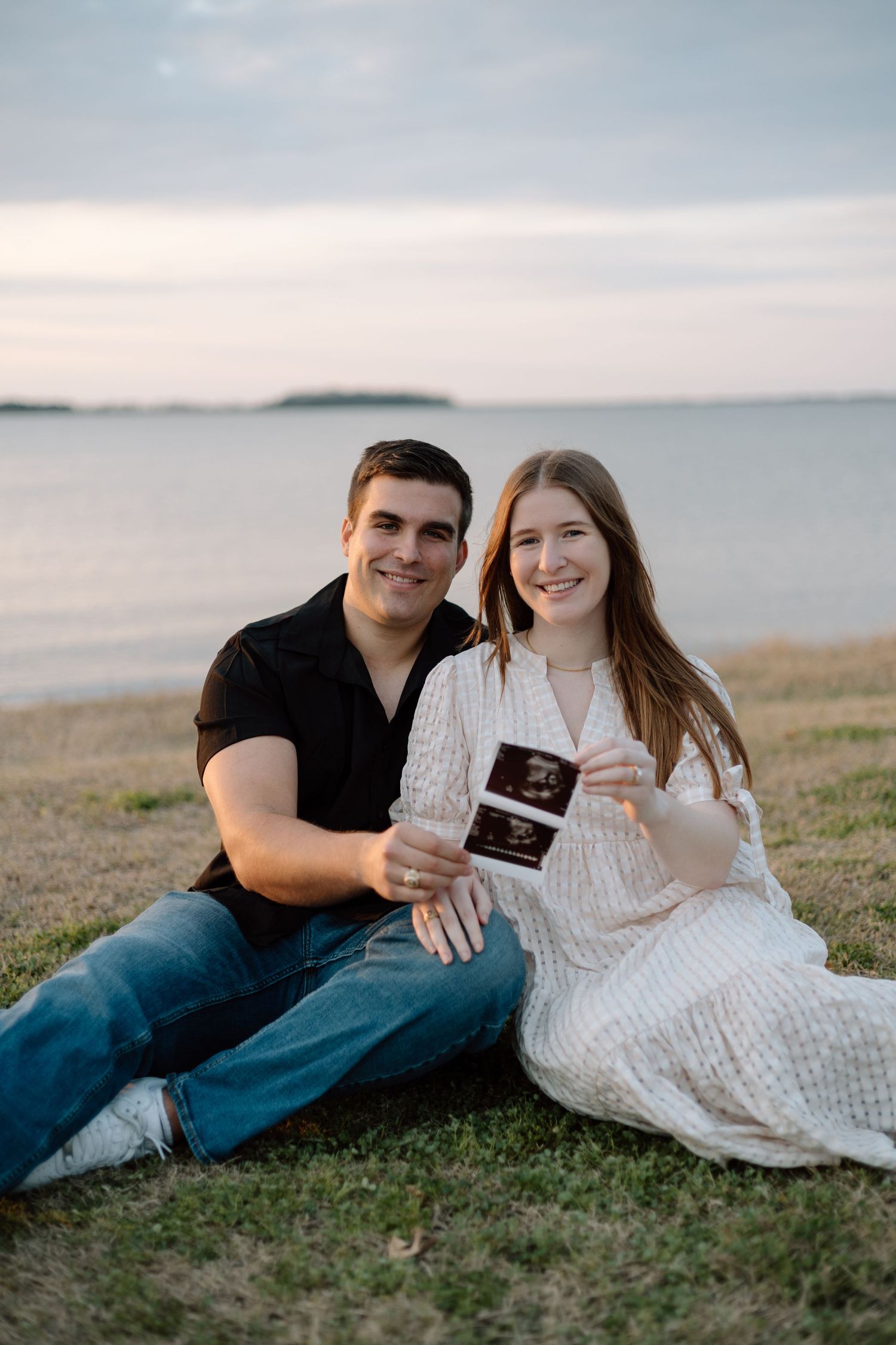 Couple sits on grass by water holding ultrasound photo in sunset lighting.