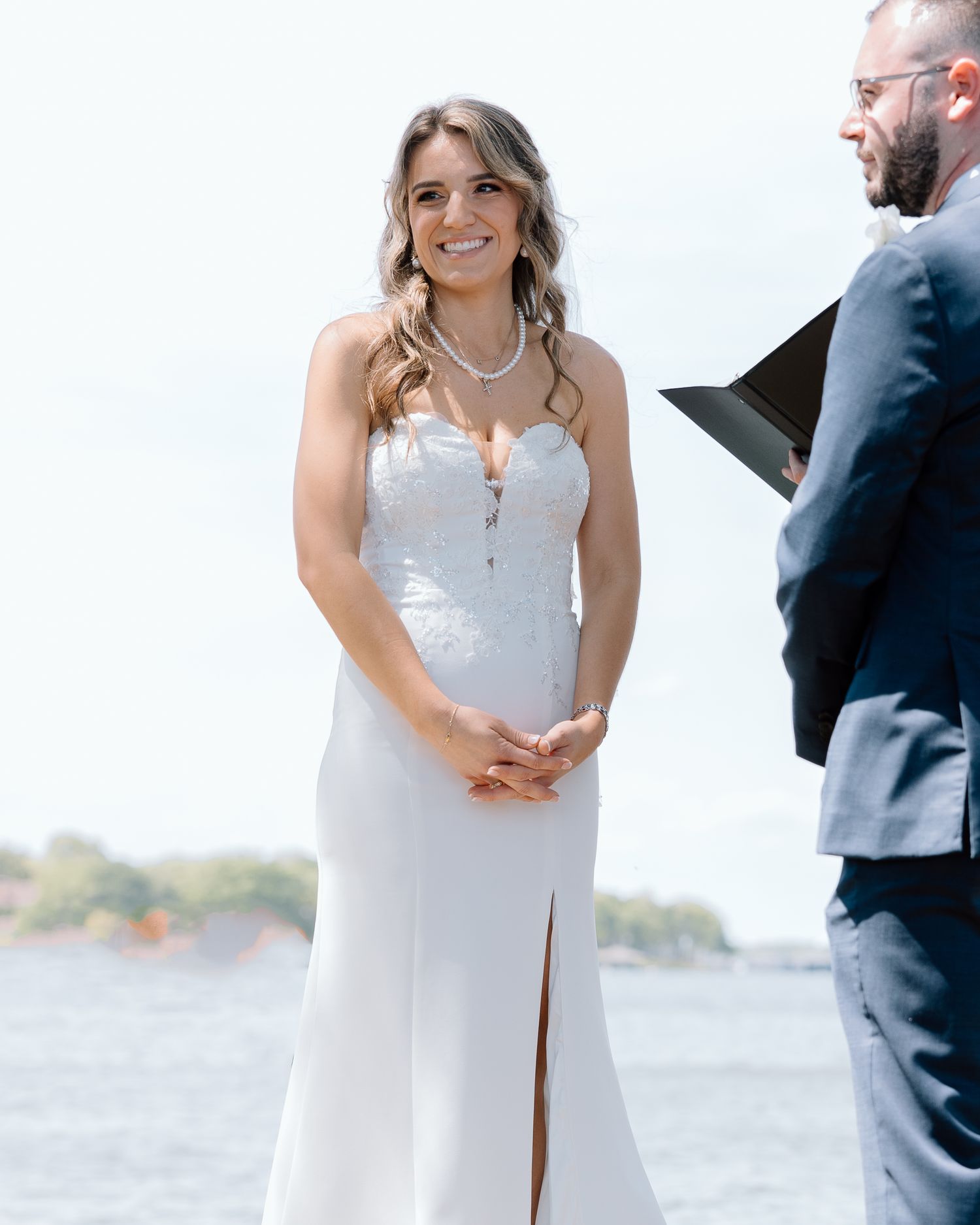 A bride in a white wedding dress stands at a waterfront ceremony location.