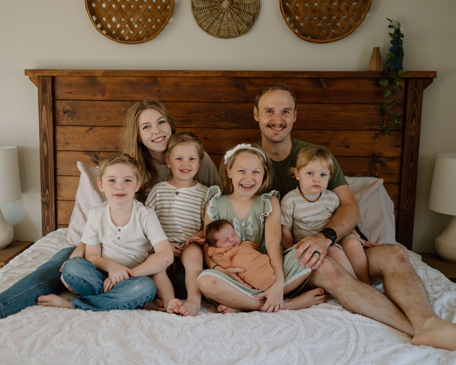 A group sits together on a white bed with decorative woven baskets hanging on the wooden headboard wall.