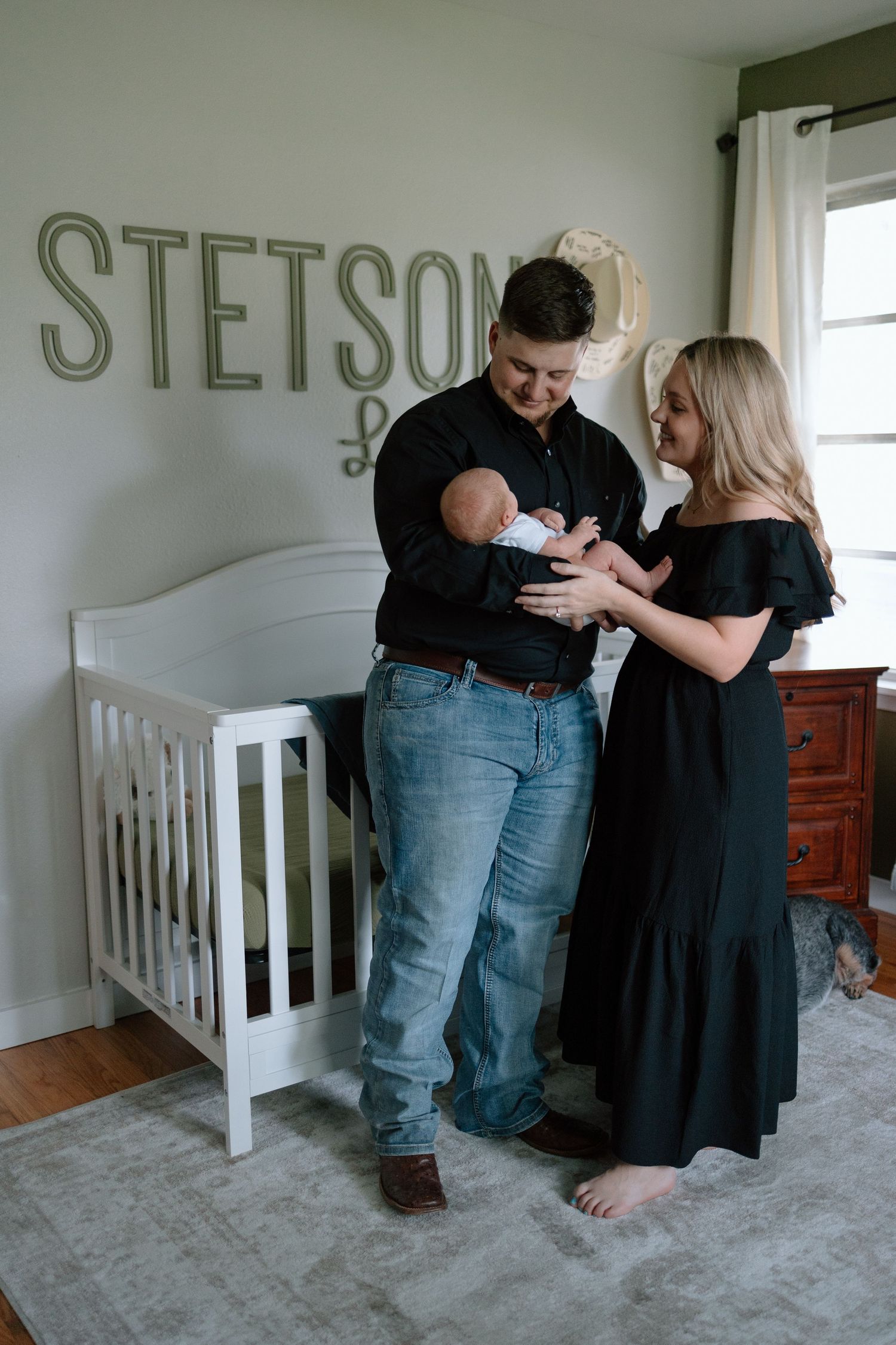 A couple tenderly holds a newborn baby in a nursery with 'STETSON' text on the wall and a white crib.