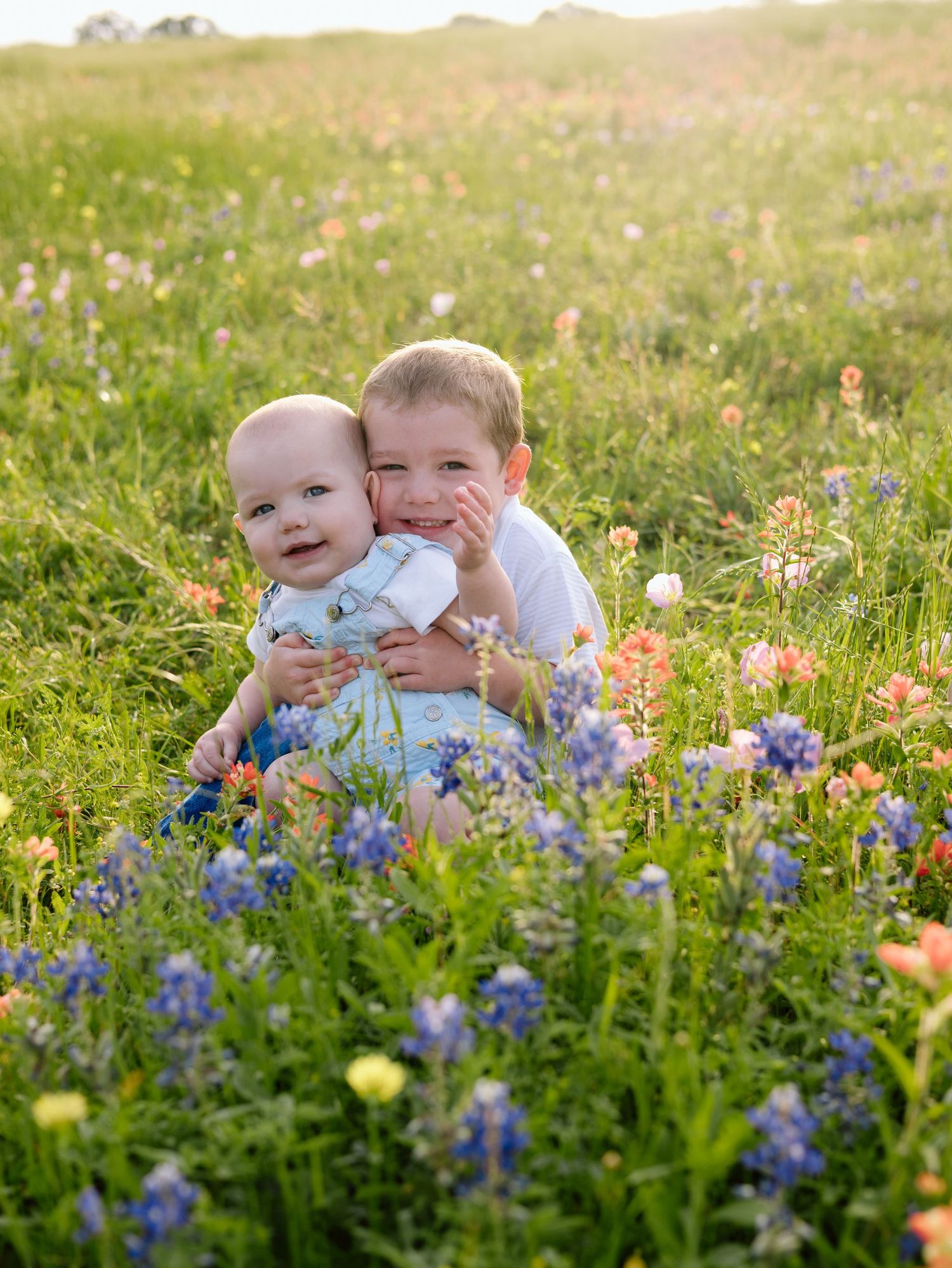 Two children sitting together in a meadow of vibrant wildflowers including bluebonnets on a sunny spring day.