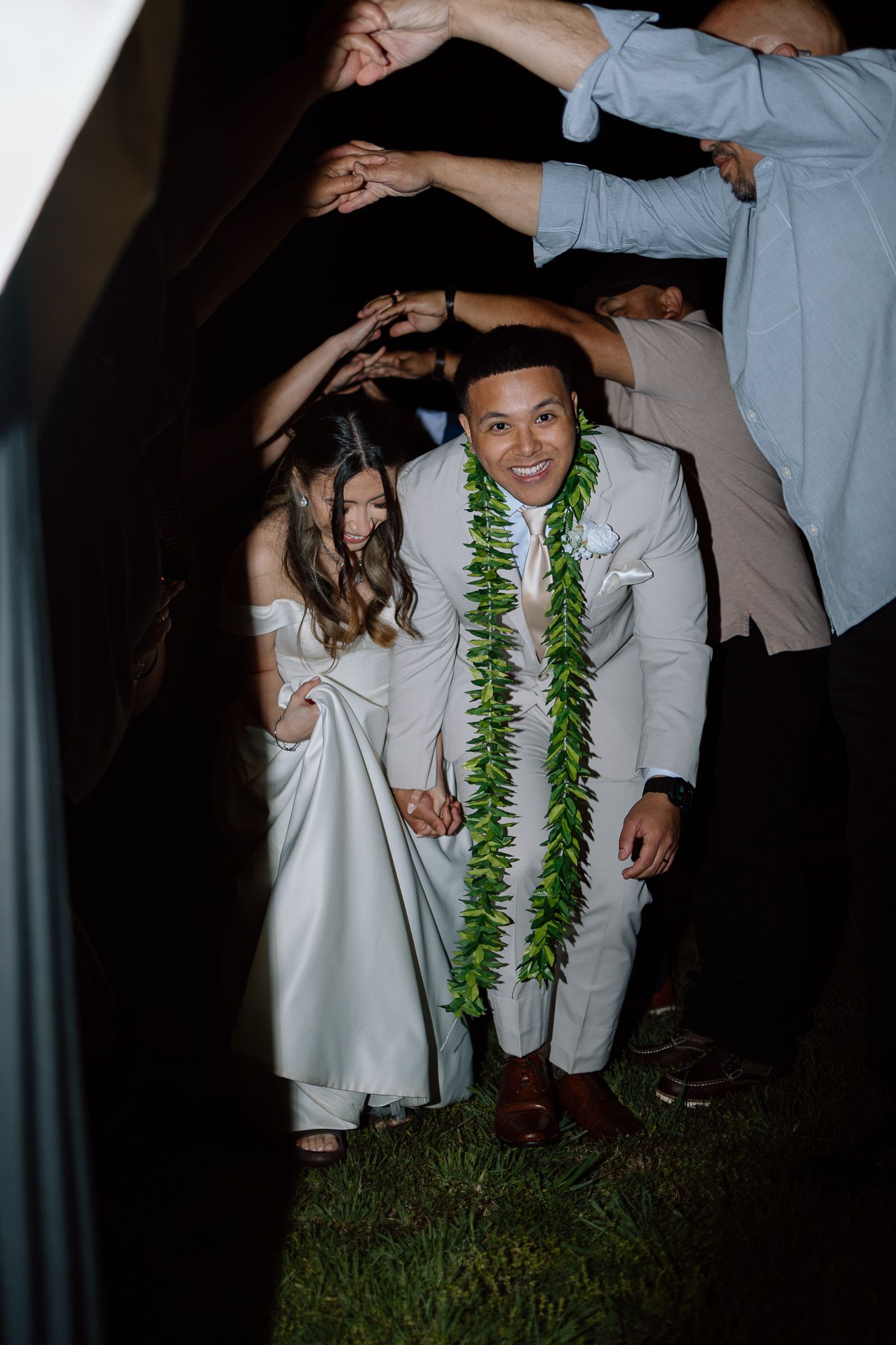 Bride and groom pass under tunnel of raised arms during wedding reception with groom wearing traditional Hawaiian lei.