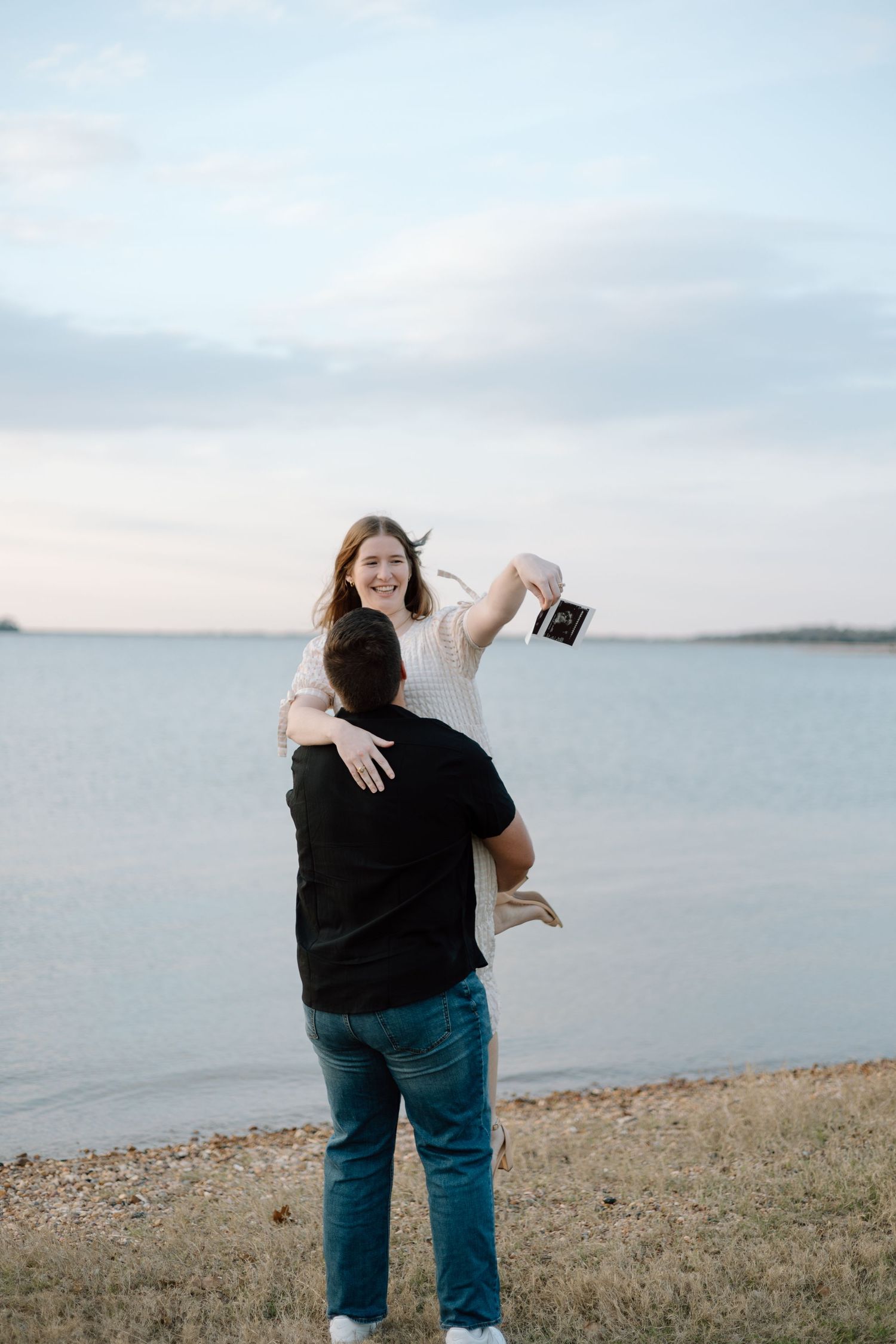 A couple embraces playfully by a waterfront at sunset as one person lifts the other in celebration.
