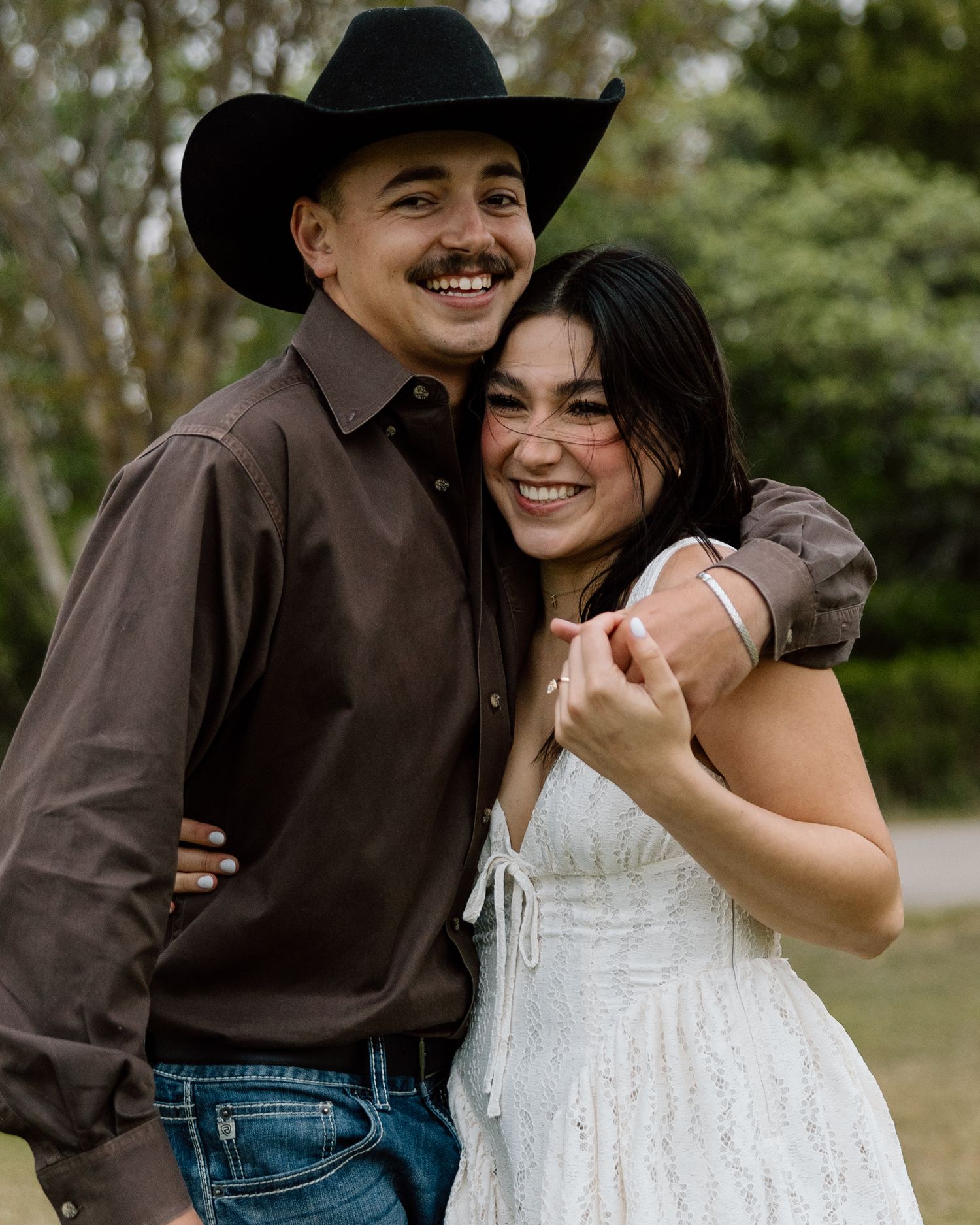 Smiling couple posing outdoors, man in black cowboy hat and brown shirt, woman in white dress, embracing happily.