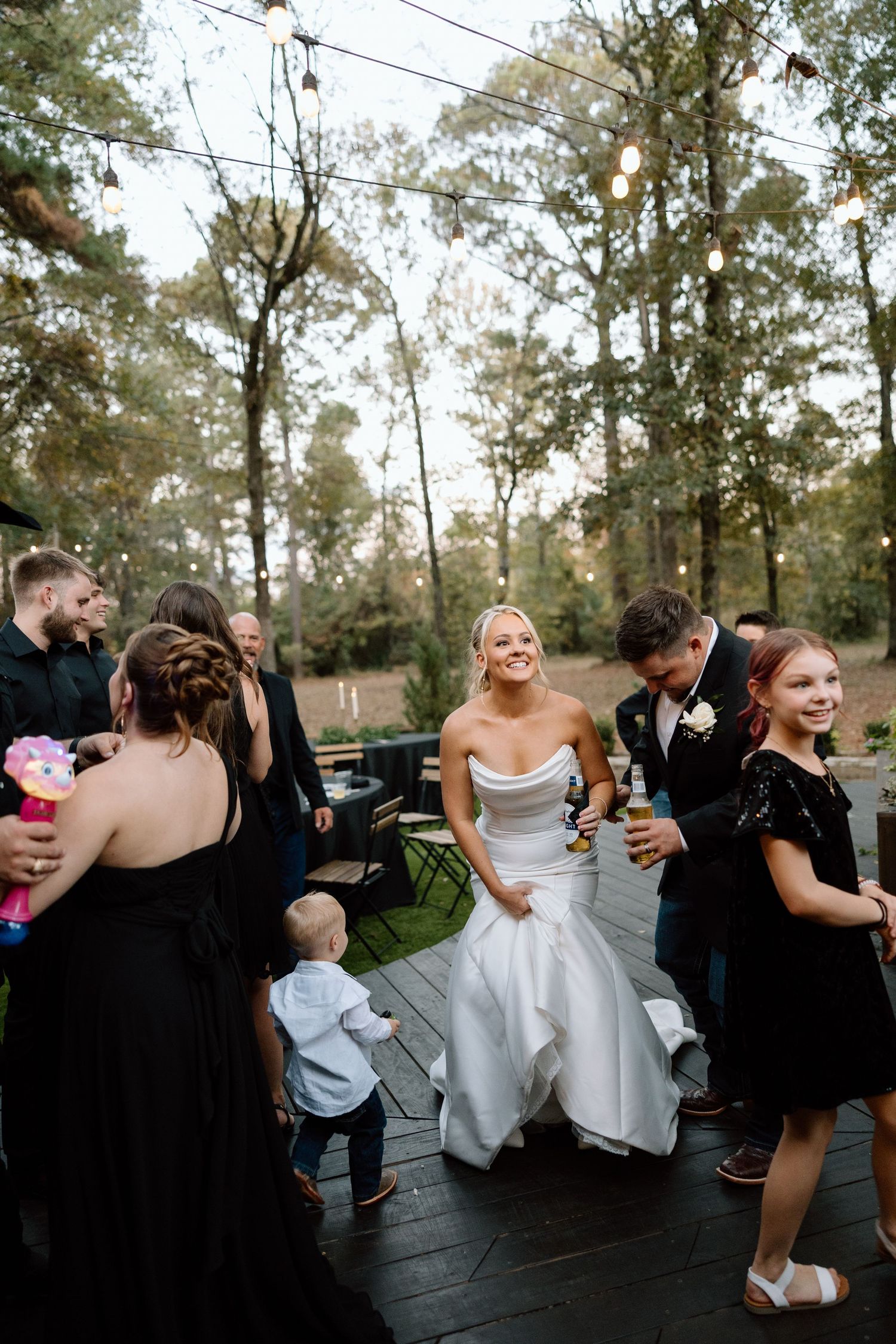 Bride in white gown laughs with guests on outdoor deck at woodland wedding reception with string lights.