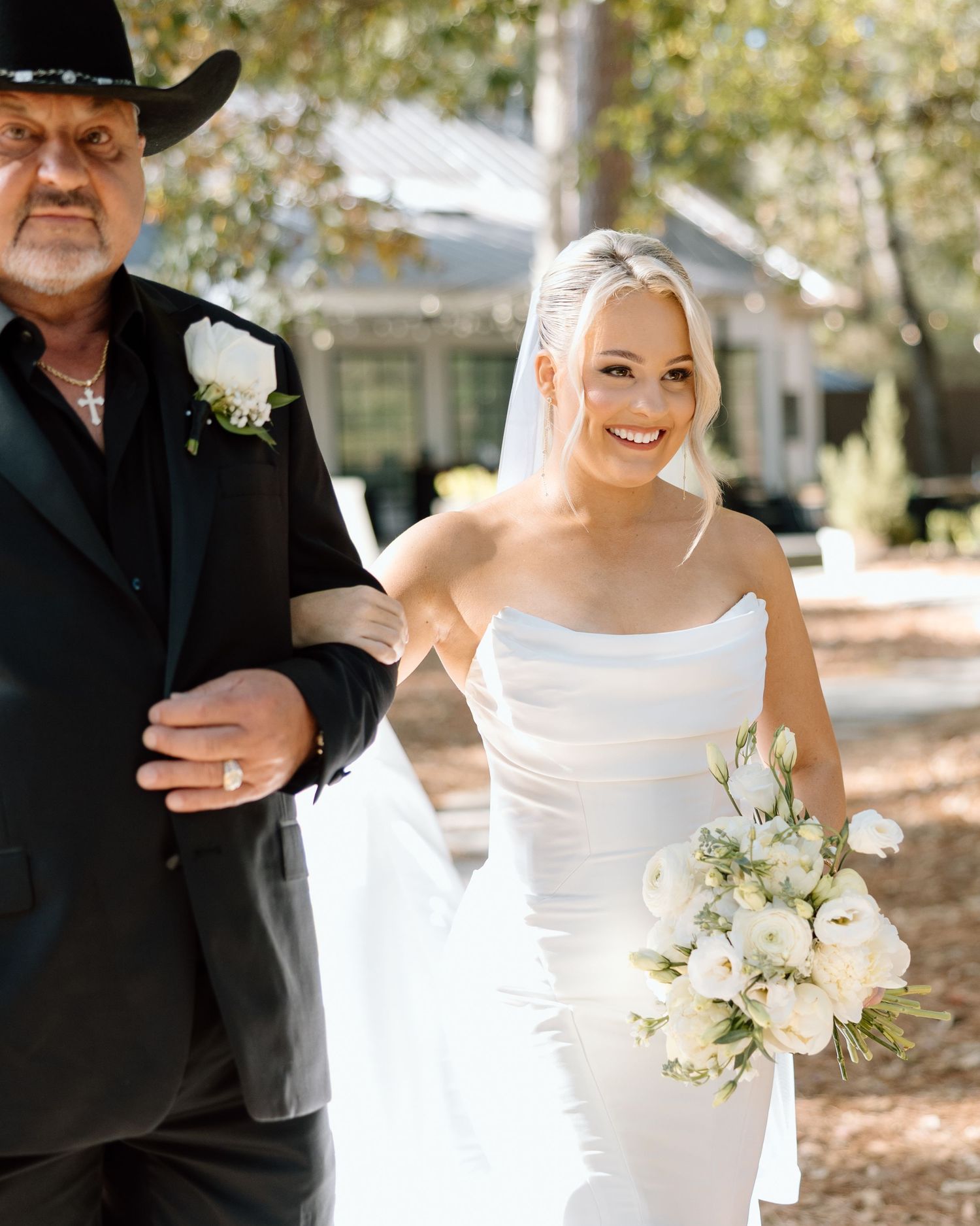 Bride in white strapless gown holding white floral bouquet walks with older man in black suit and cowboy hat outdoors.
