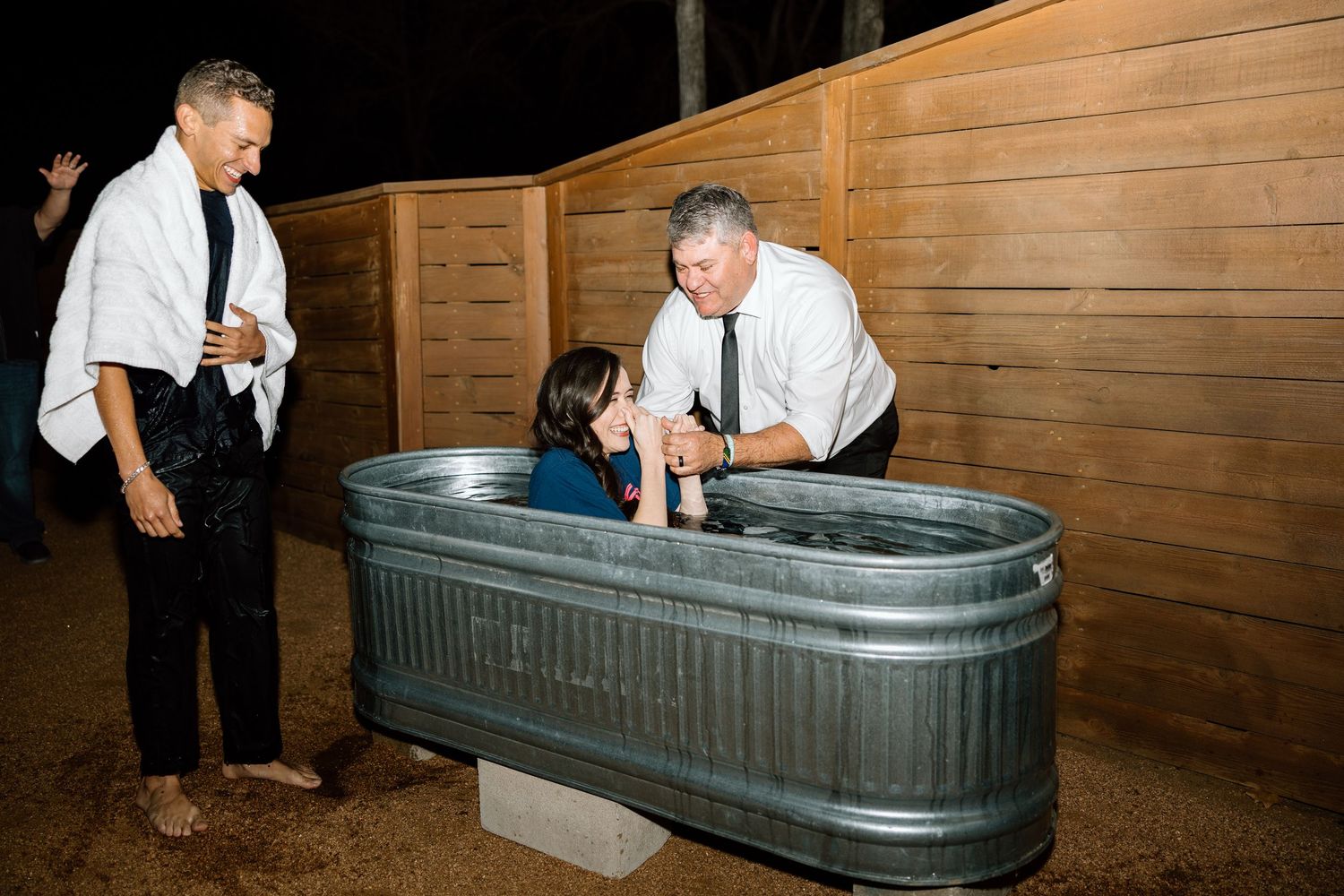 A child being baptized in a metal trough tub outdoors at night with two men assisting.