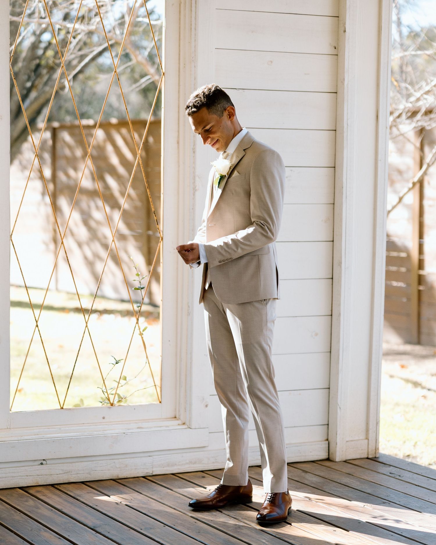 Groom in tan suit standing on wooden porch beside decorative glass door.