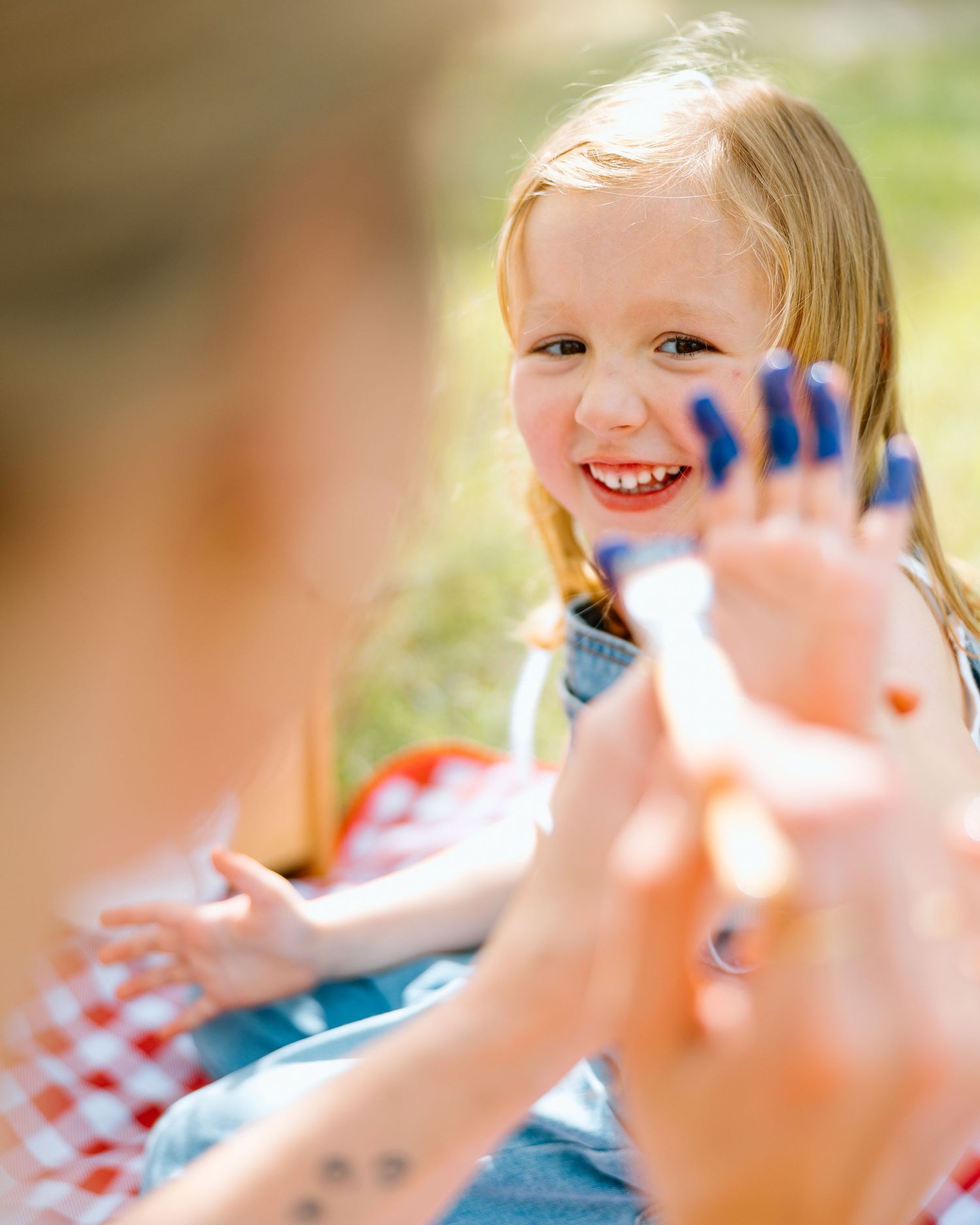 A cheerful blonde girl showing blue paint-covered fingers to an adult during an outdoor painting activity.