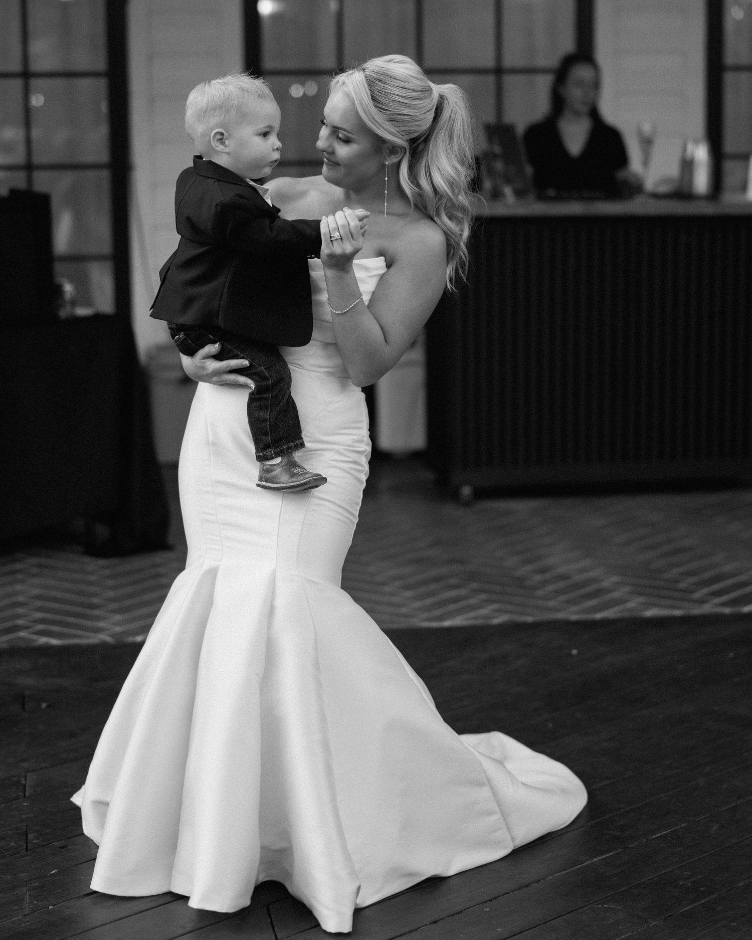 Bride in mermaid gown dances with young boy in tuxedo at wedding reception.
