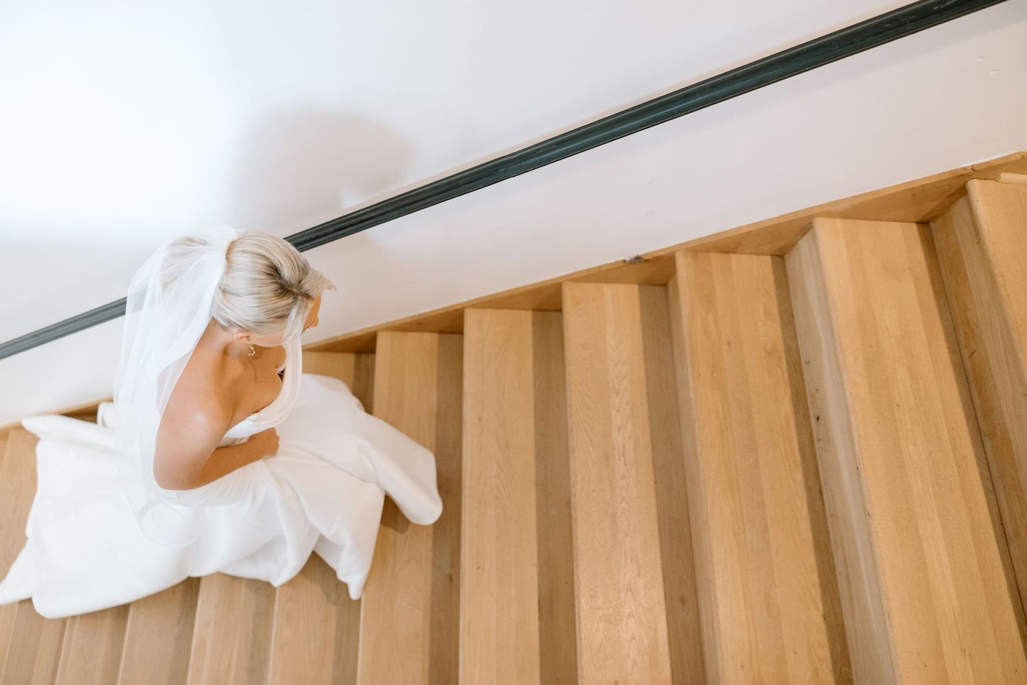 Bride in white dress and veil sitting on modern wooden staircase, viewed from above.