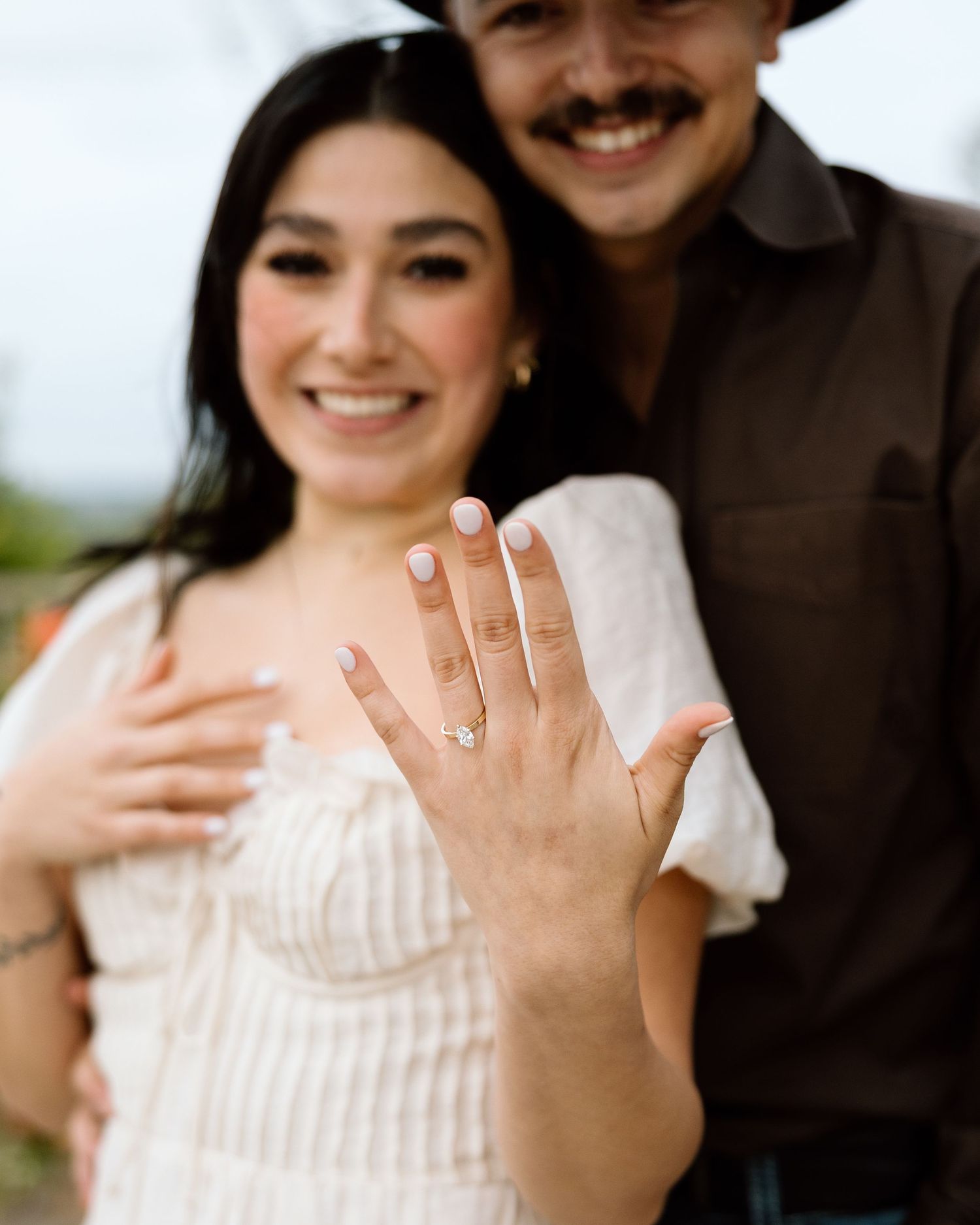 Smiling couple showing off engagement ring, woman in white dress extending hand toward camera outdoors.