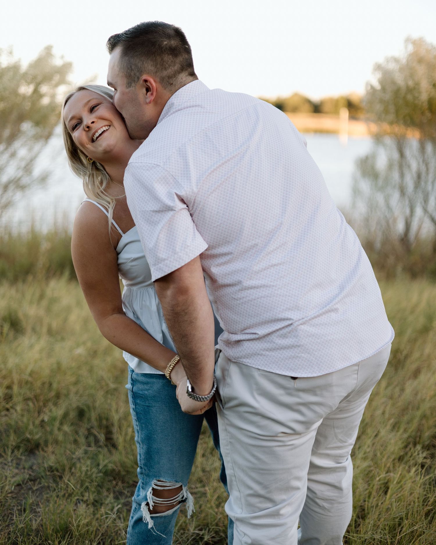 A couple sharing a romantic moment outdoors near a lake, man kissing woman's cheek as she laughs joyfully.