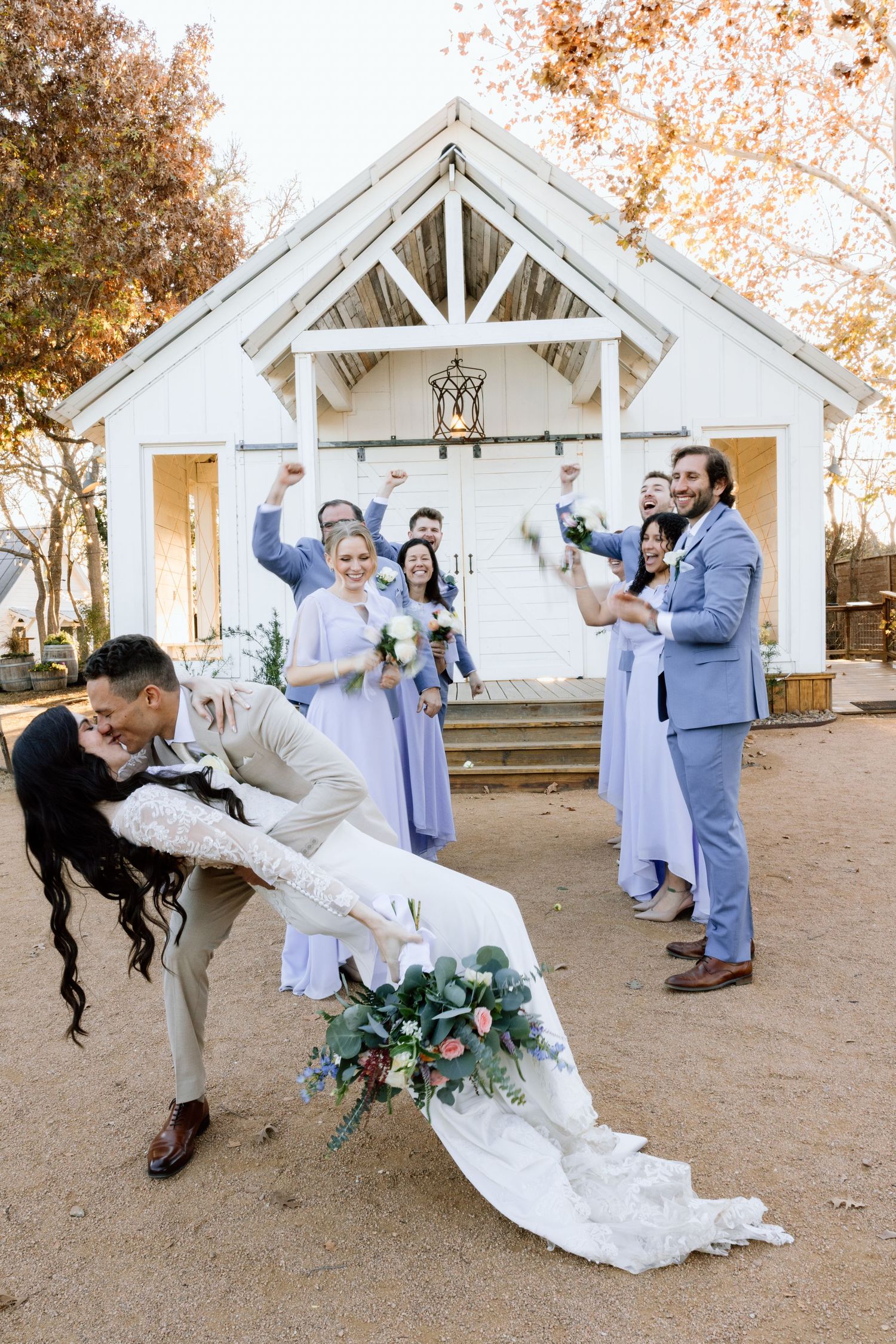 Groom dips bride for kiss outside white chapel as wedding party in lavender attire cheers behind them.