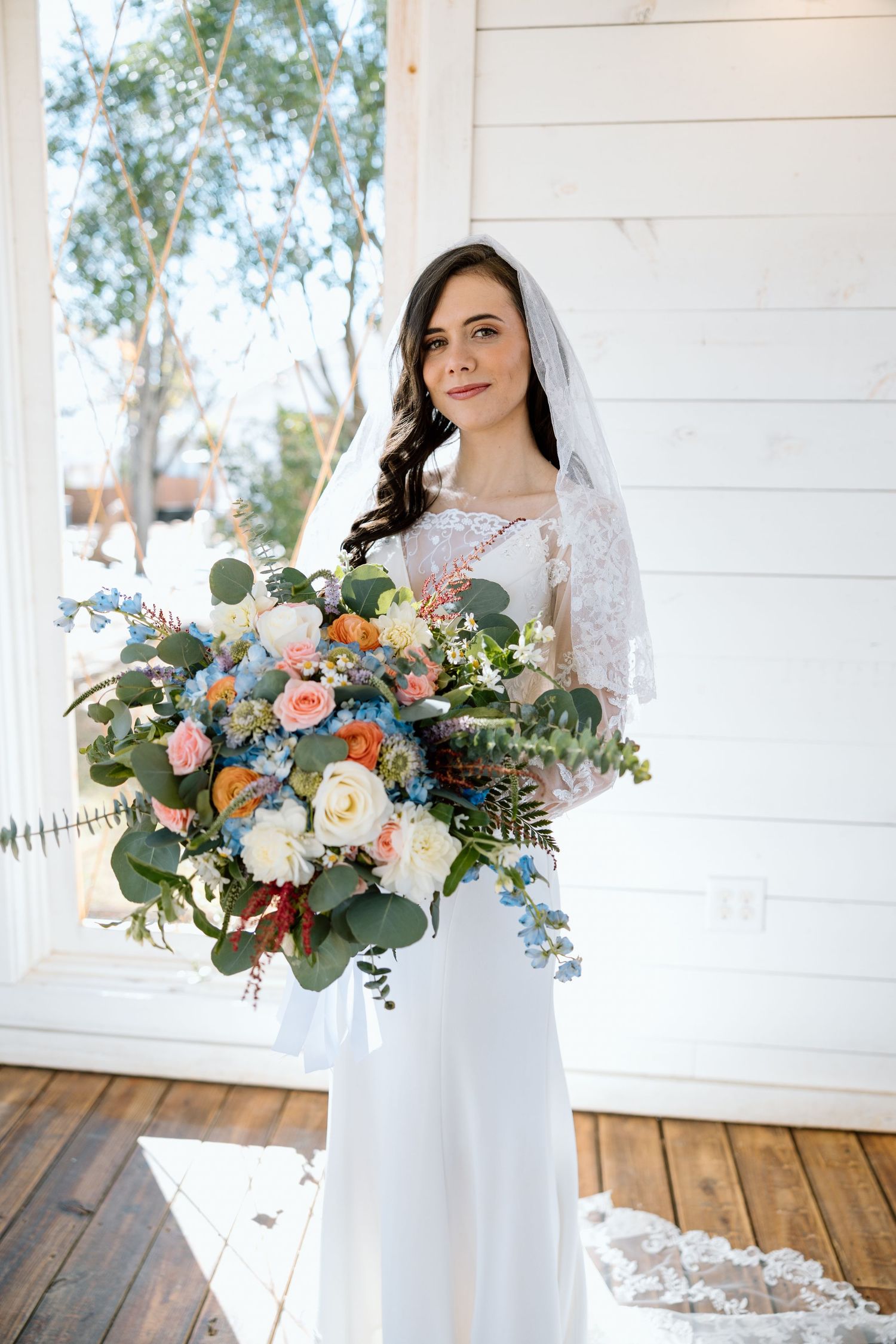 Brunette bride in white lace gown holding large colorful bouquet of roses and eucalyptus.