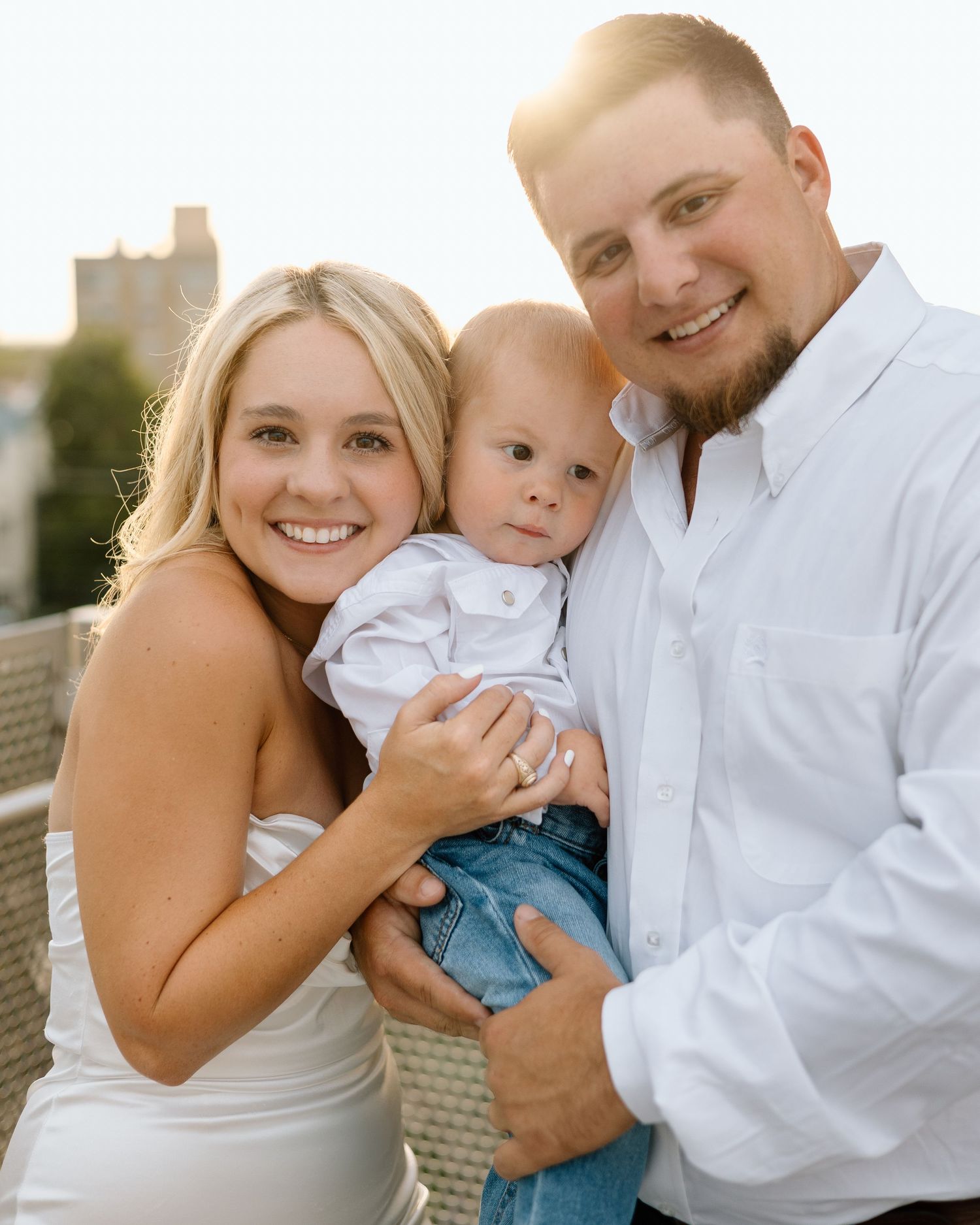 A smiling young family with a blonde woman, bearded man in white shirt, and baby boy posing outdoors at golden hour.
