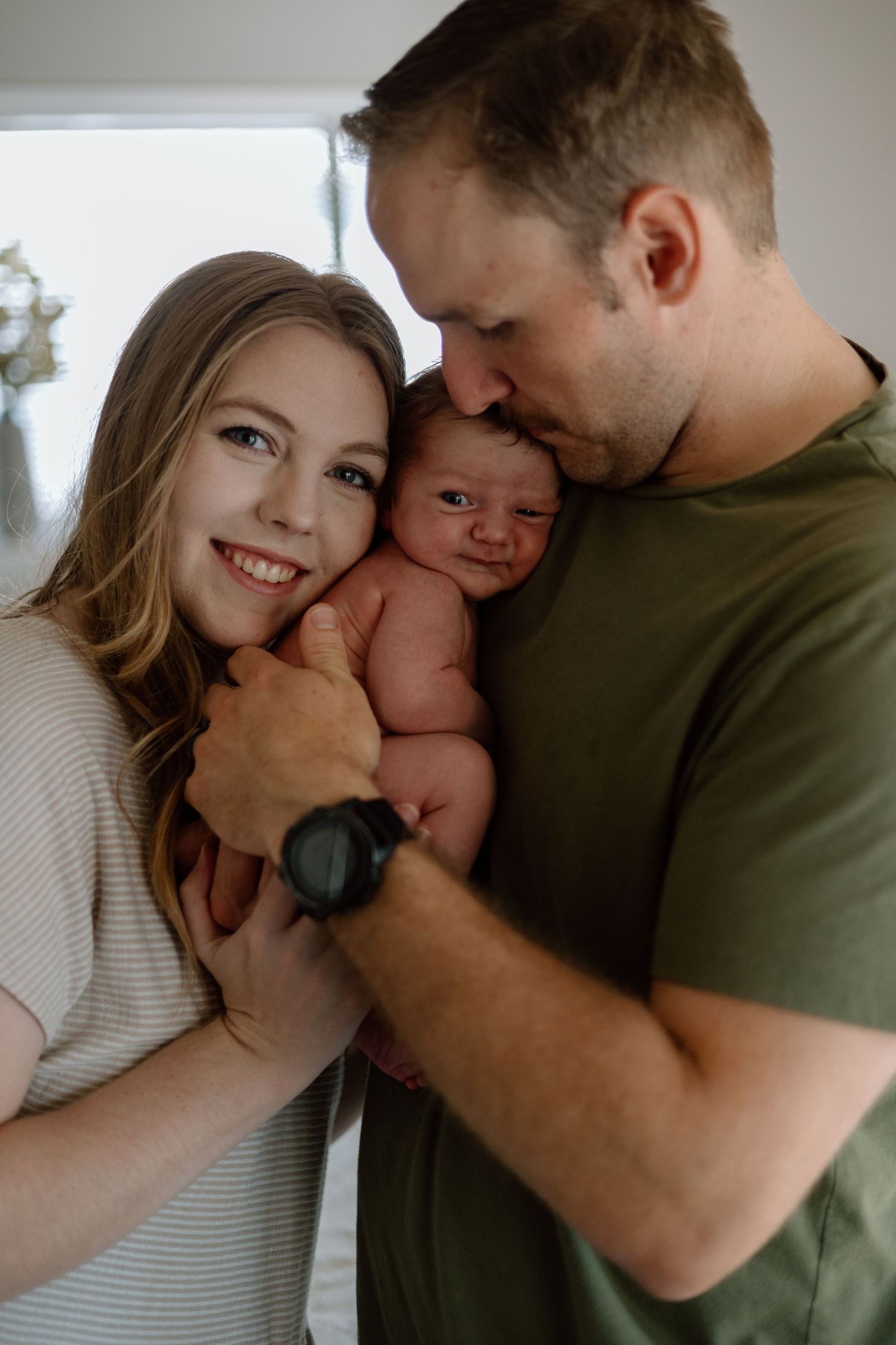 An intimate family portrait shows parents lovingly snuggling their newborn baby in a tender indoor moment.