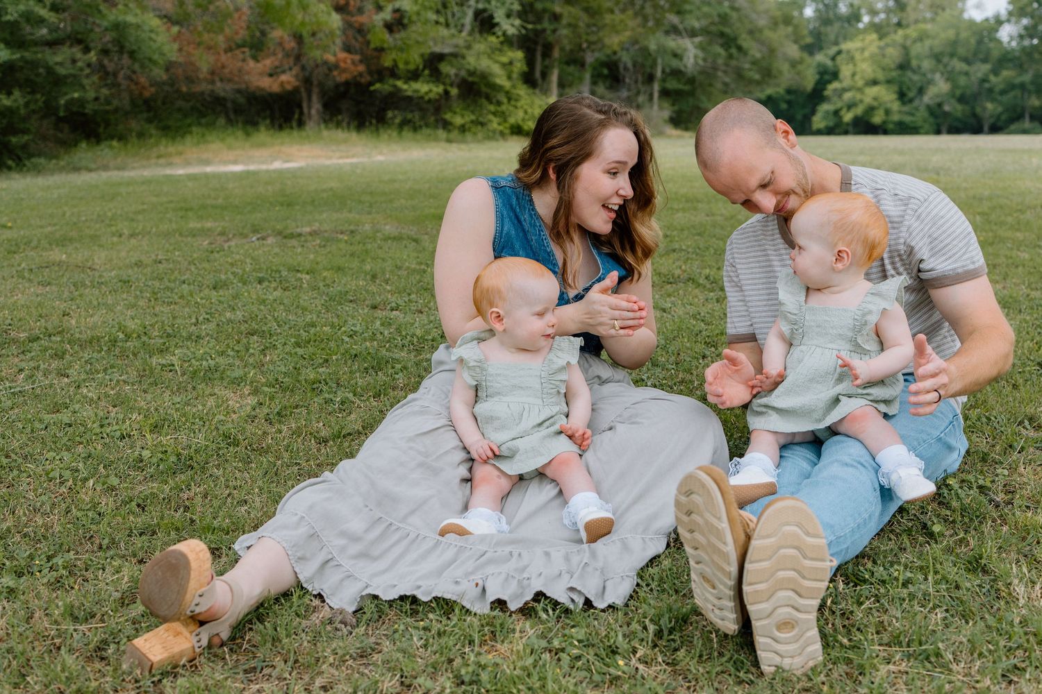 A family sits together on a blanket in a grassy field on a sunny day, sharing a joyful moment outdoors.