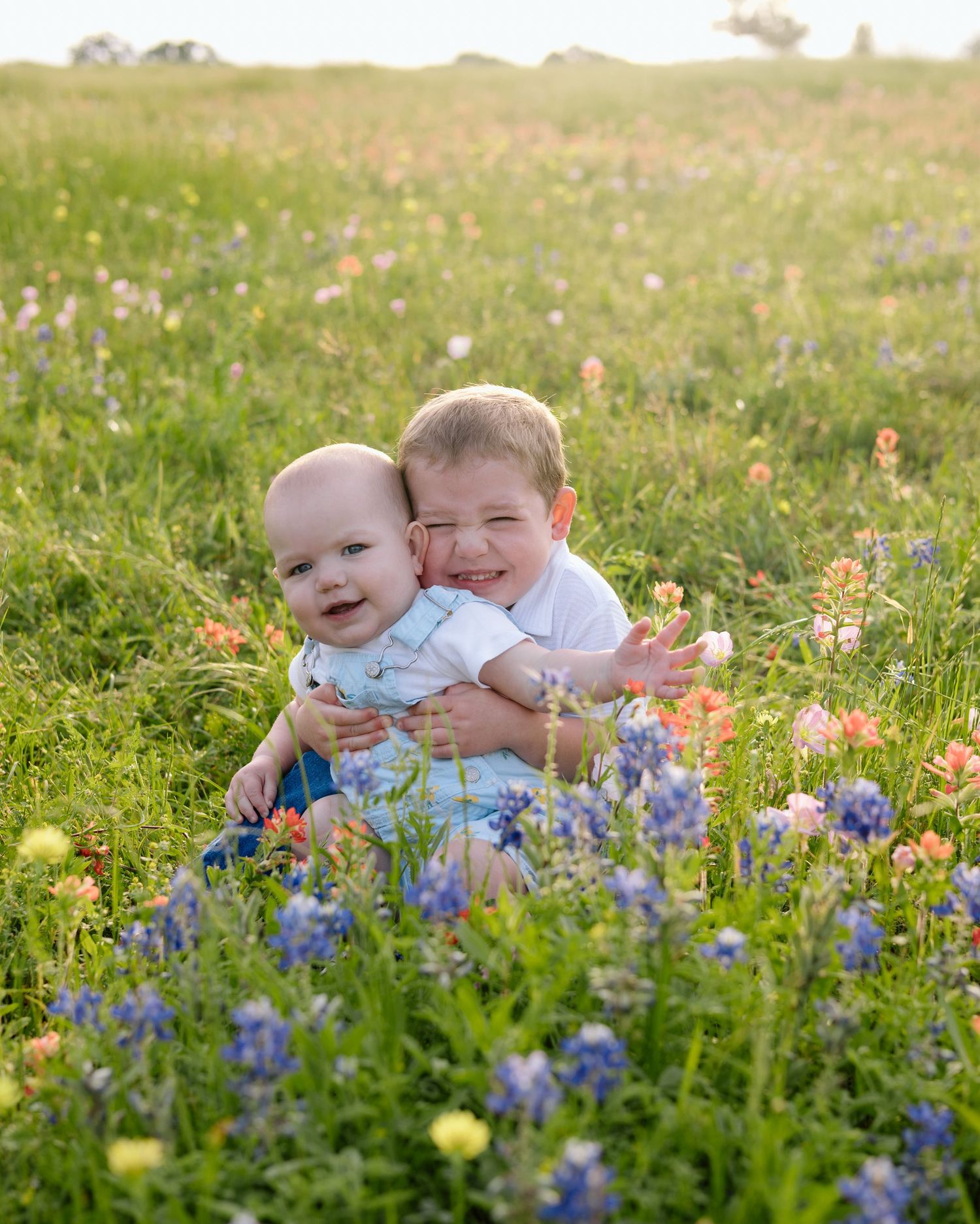 Two children sit together in a meadow with vibrant blue wildflowers and yellow blooms on a sunny day.