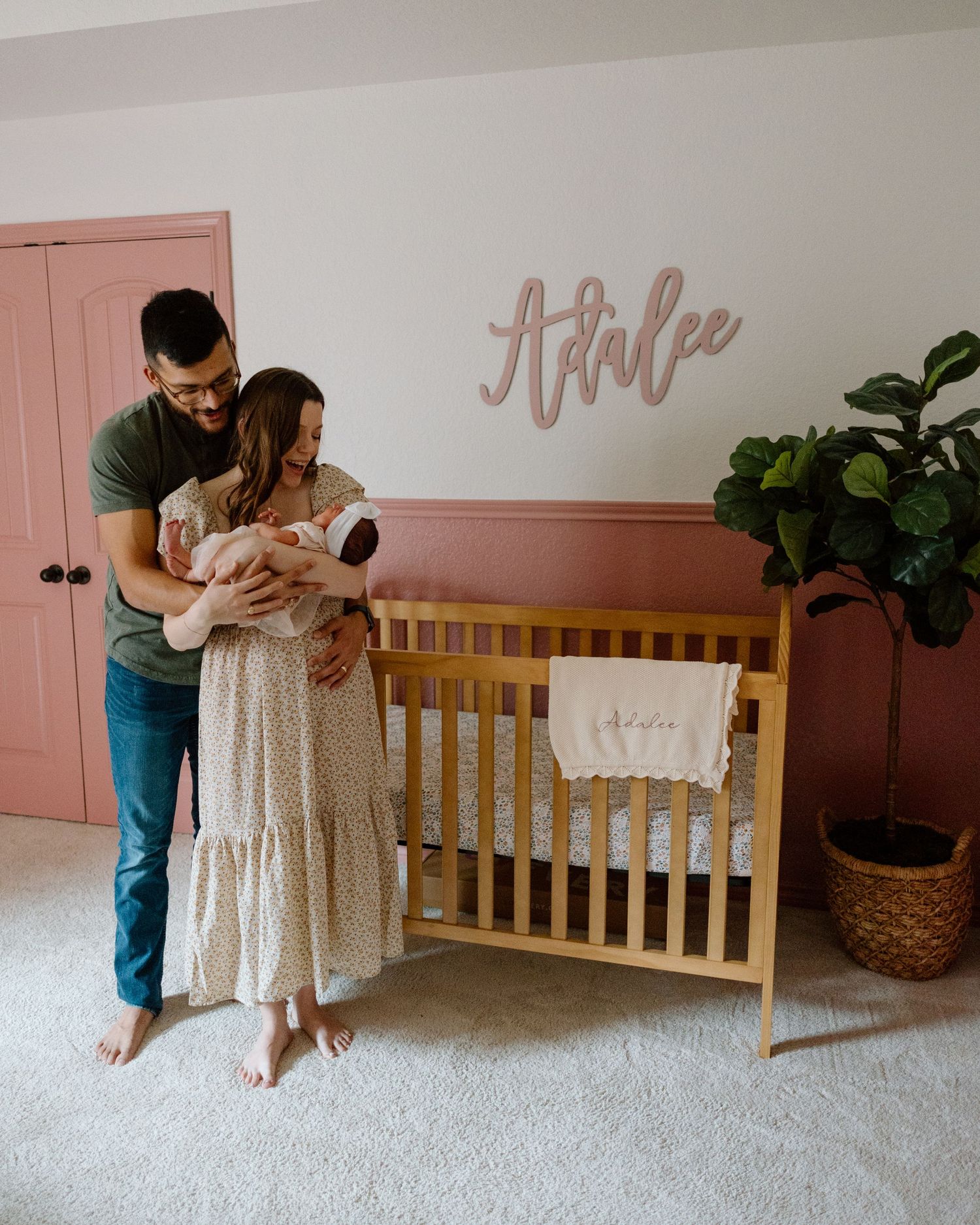 A nursery scene with wooden crib, pink accent wall, and name decoration in rose gold lettering.