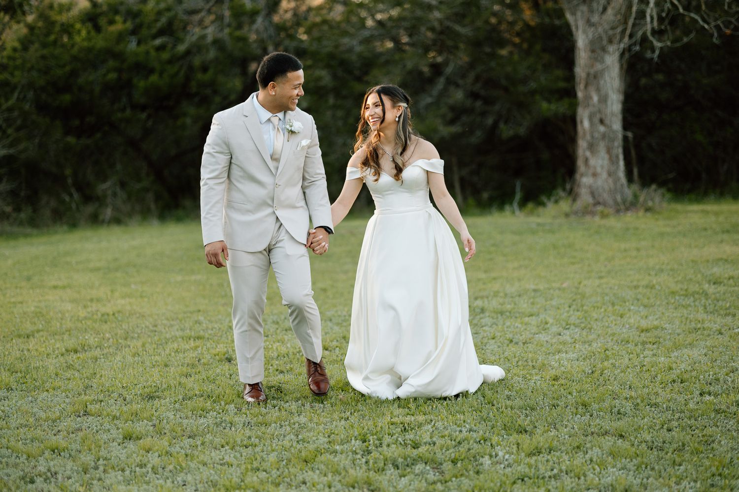 Happy couple laughing together on grass while wearing light-colored wedding attire.