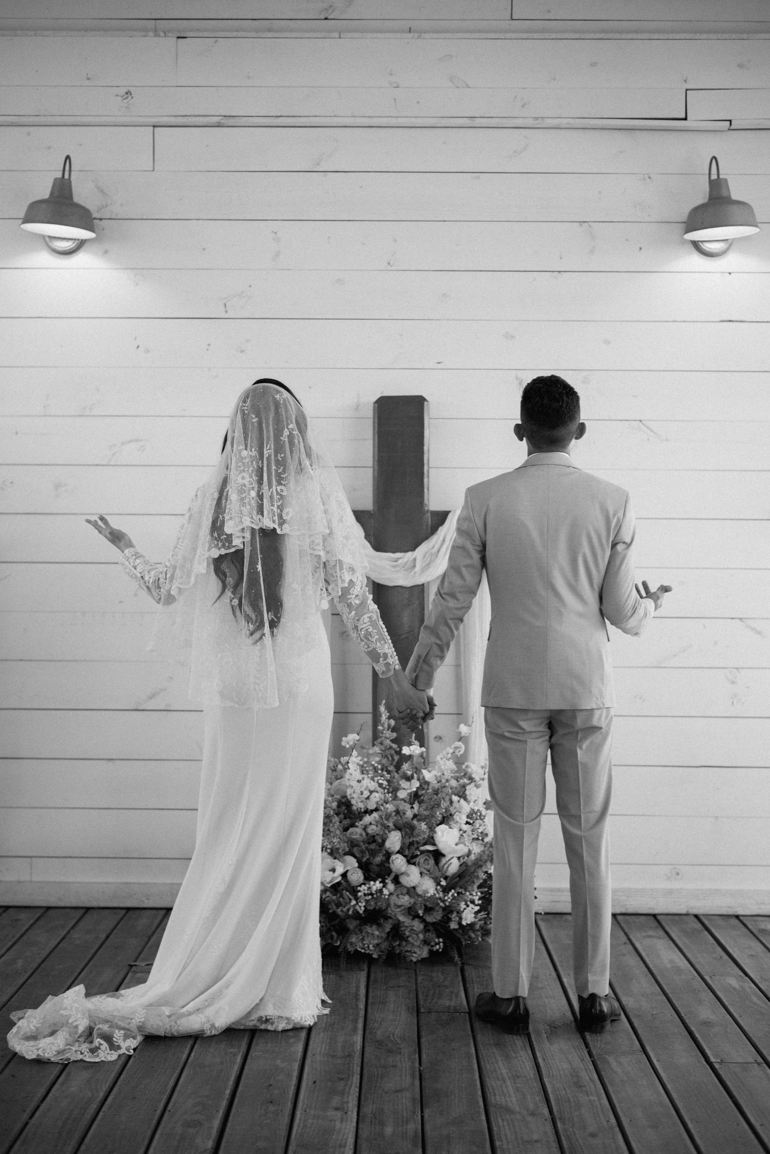Black and white photo of bride and groom holding hands in prayer before a cross at wedding.