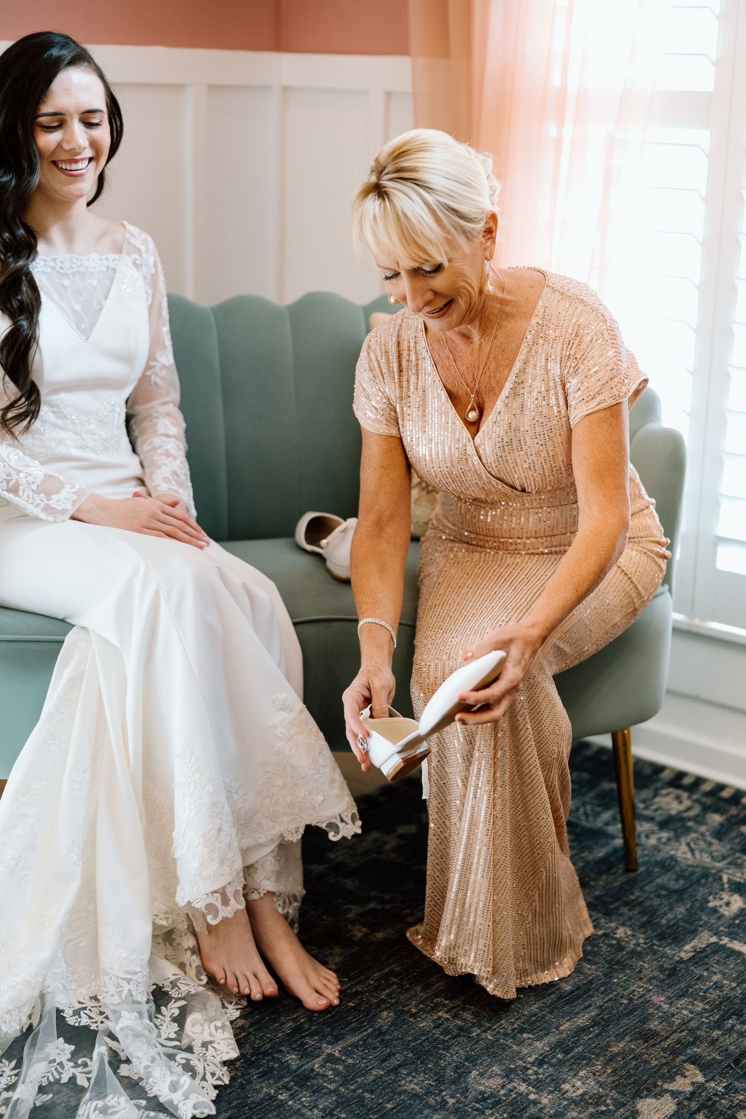 Mother of the bride helping bride put on wedding shoes while sitting on a teal sofa.