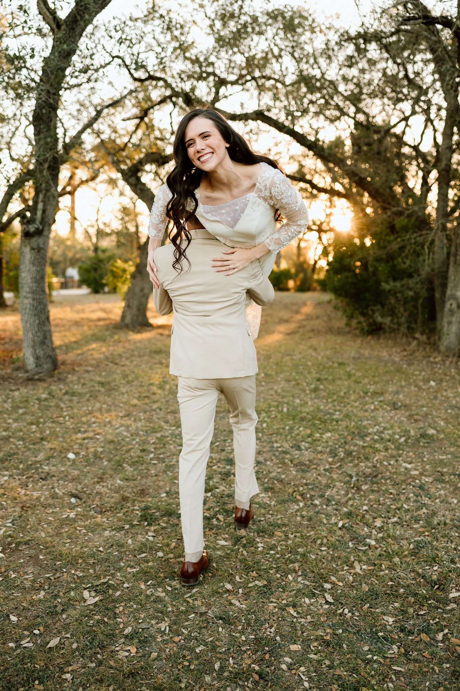 Smiling woman in beige outfit poses outdoors among trees at golden hour sunset.