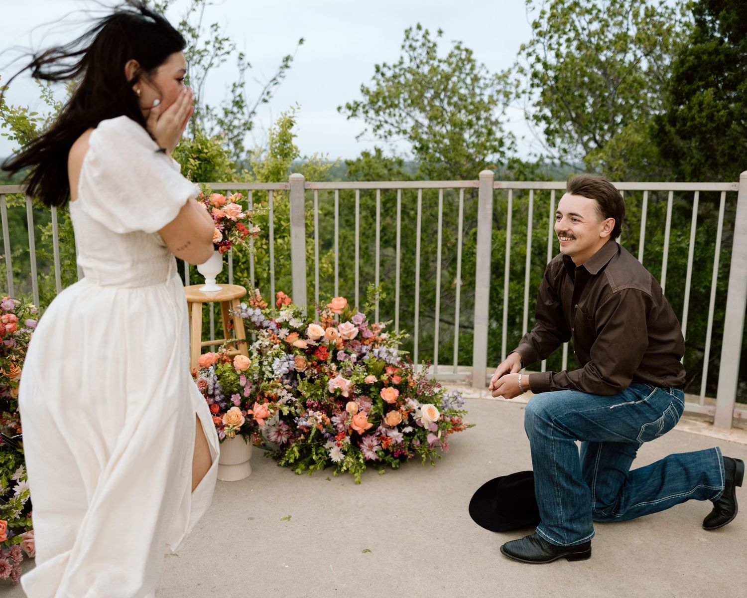 Man on one knee proposing to woman in white dress on a balcony with floral arrangement.